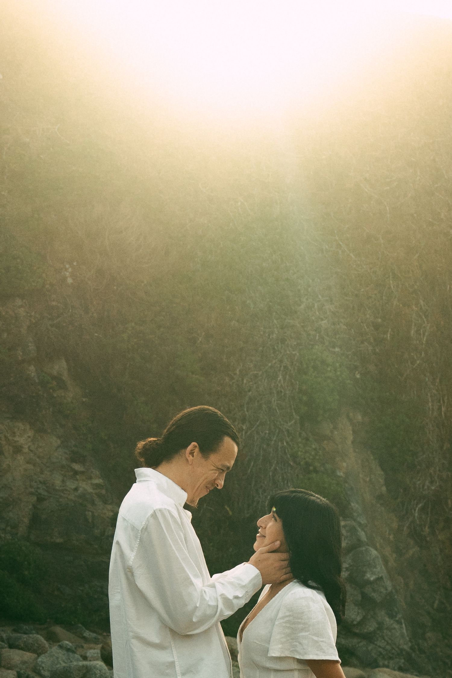 A couple in white clothing share an intimate moment against a misty natural backdrop with rocky cliffs.