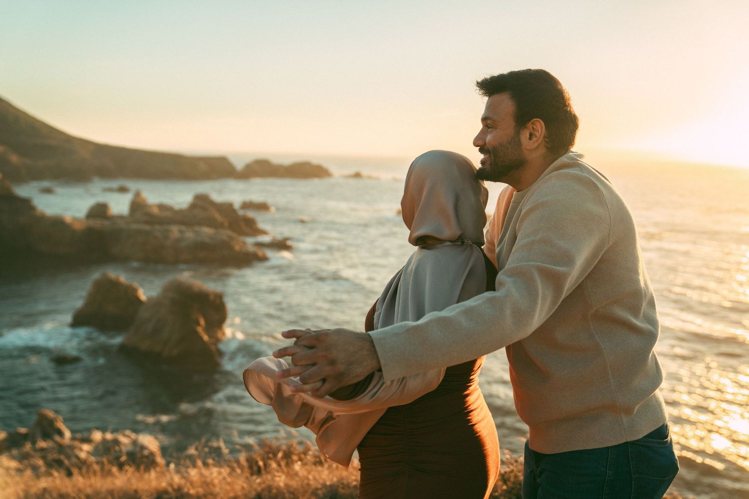 Two people share an intimate moment on rocky cliffs overlooking a golden sunset and ocean waves.