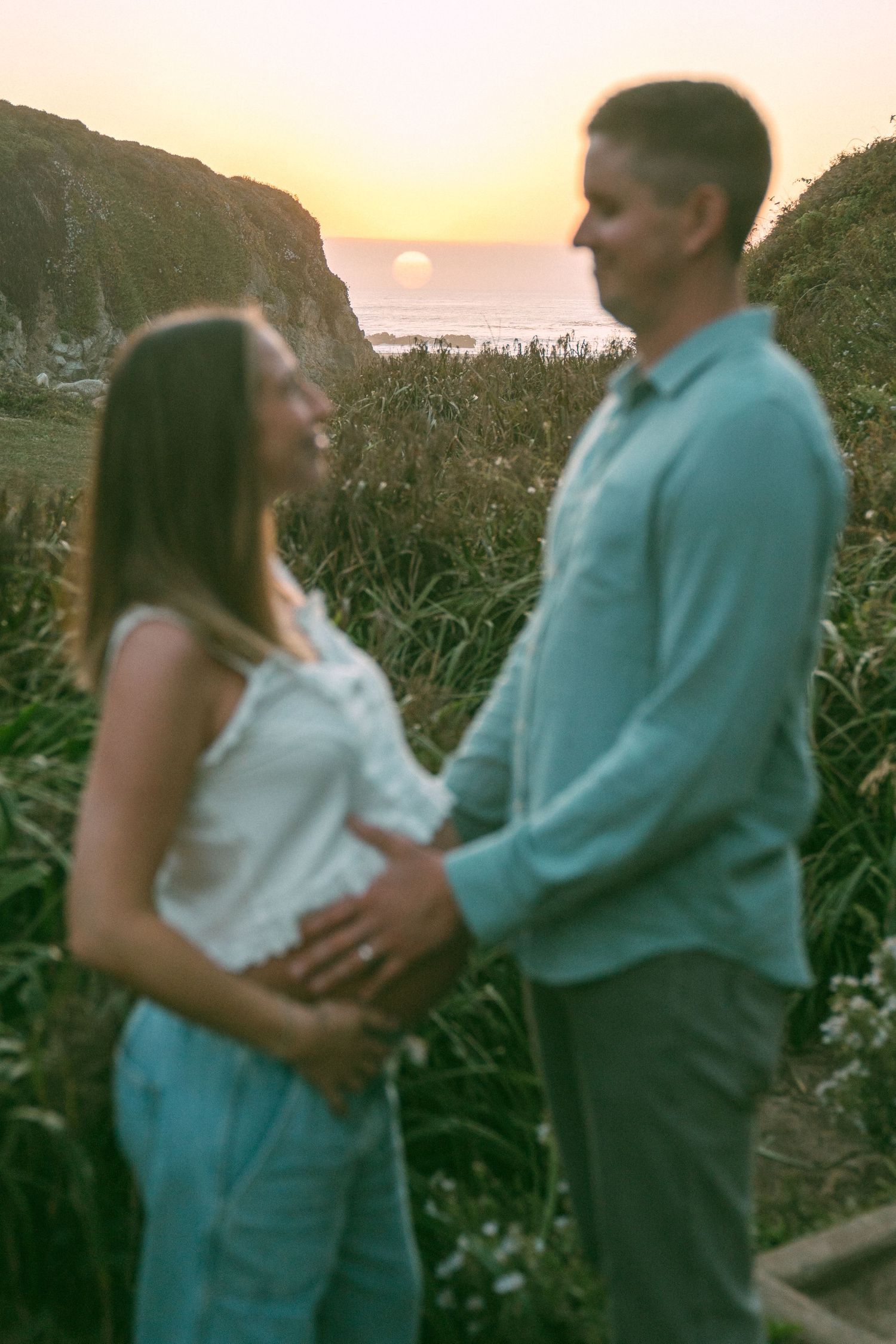 A couple embraces on a cliff overlooking the ocean at sunset among tall grass.