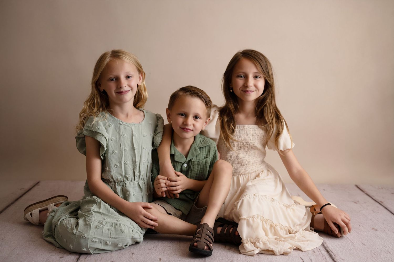 Three children siblings pose together smiling on a light wood floor against a neutral beige studio backdrop.