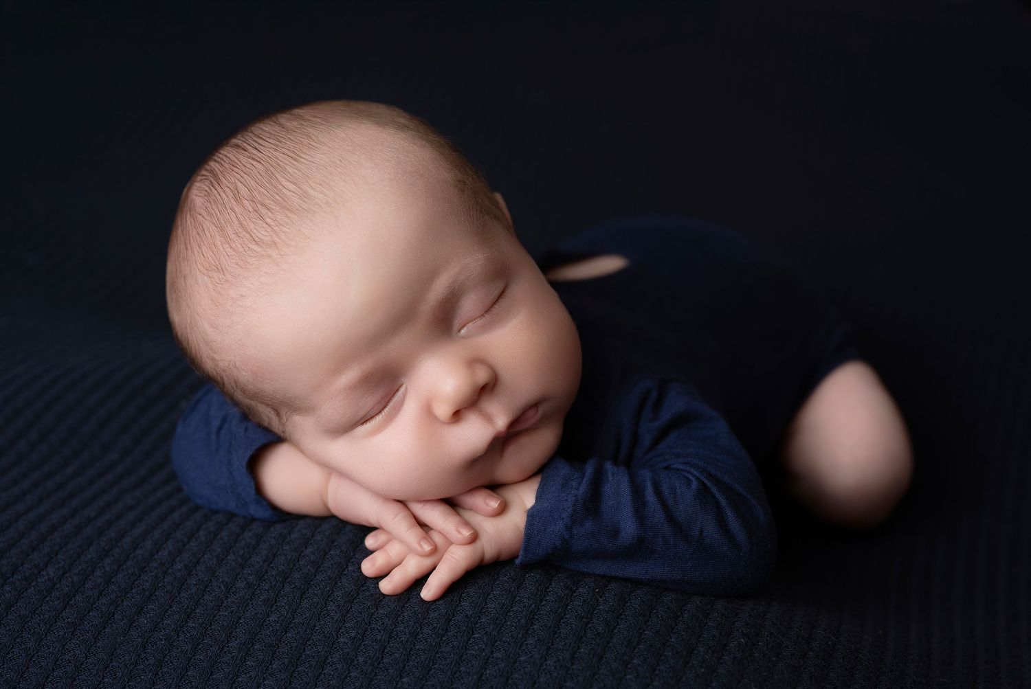 Sleeping newborn baby boy in navy outfit posed on dark blue blanket with chin on hands.