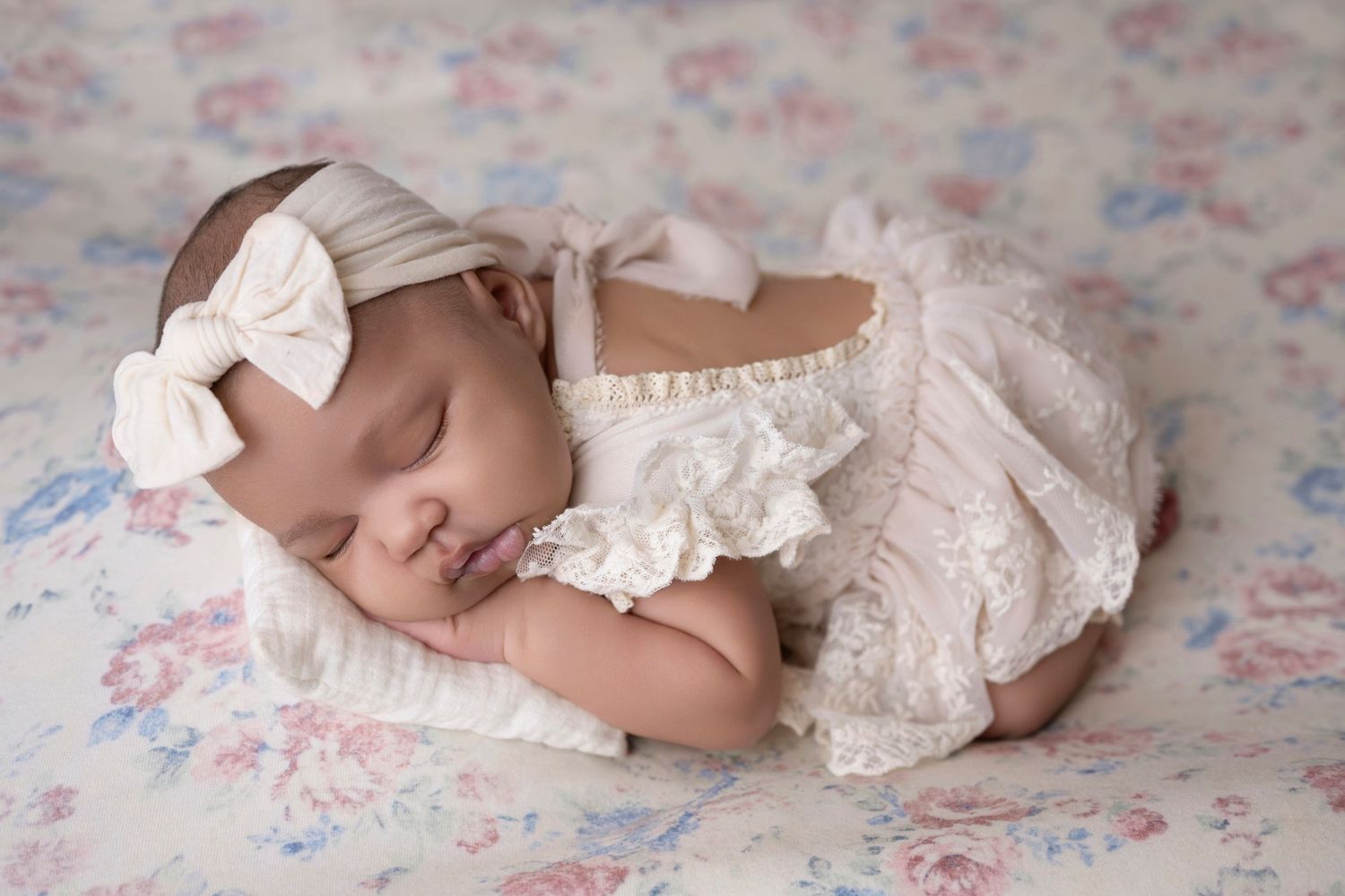 Sleeping newborn baby girl in cream lace outfit and bow headband on floral blanket.