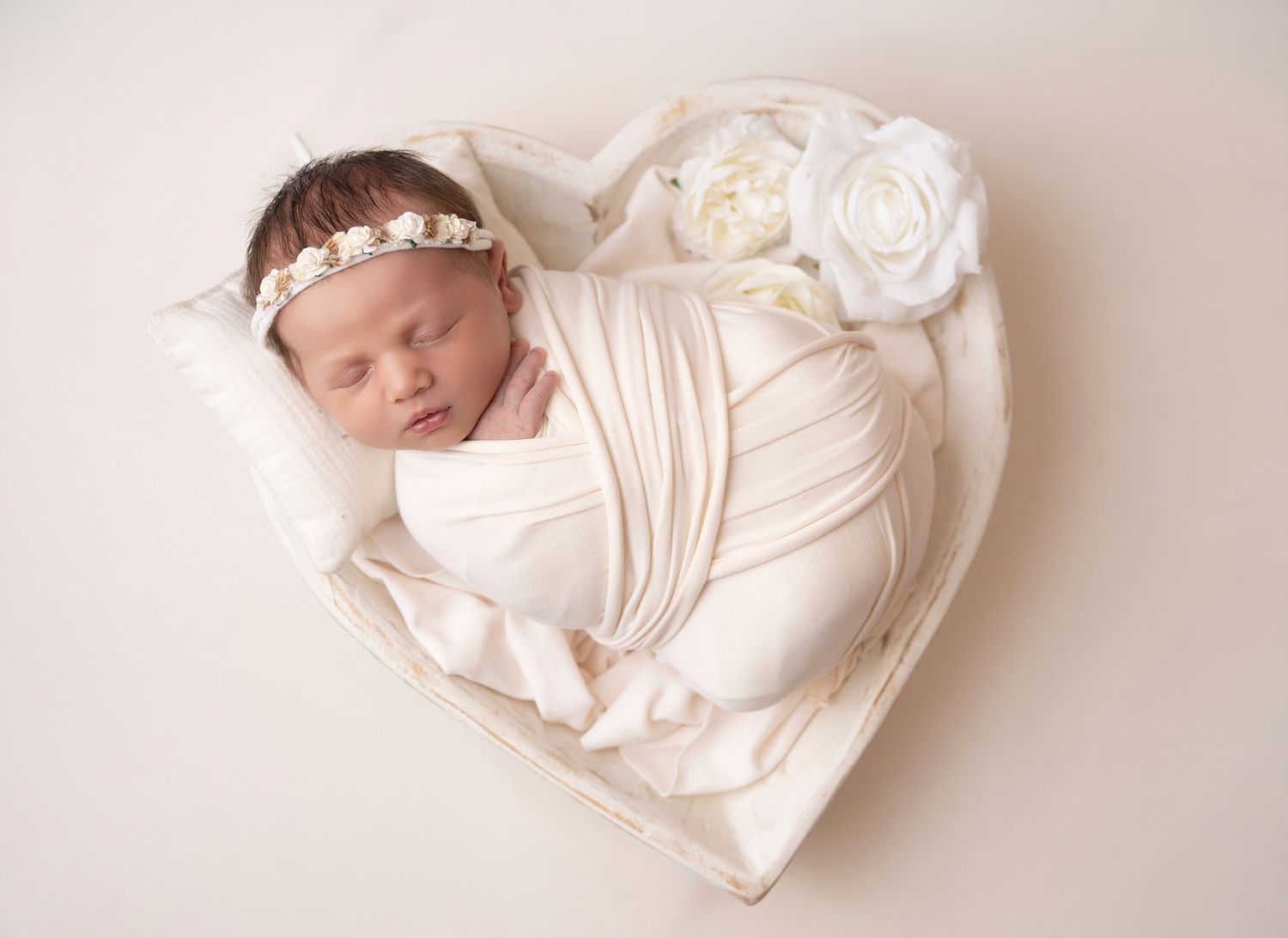 Newborn baby wrapped in white swaddle sleeping in heart-shaped prop with floral headband and white roses.