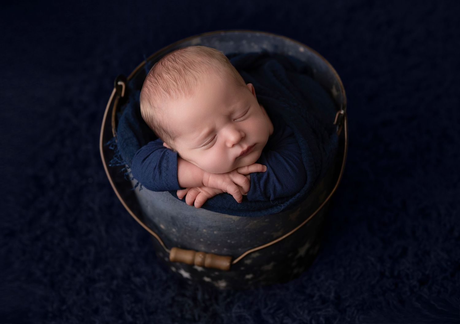 Newborn baby sleeping peacefully in a navy blue bucket with matching wrap on a dark blue textured background.