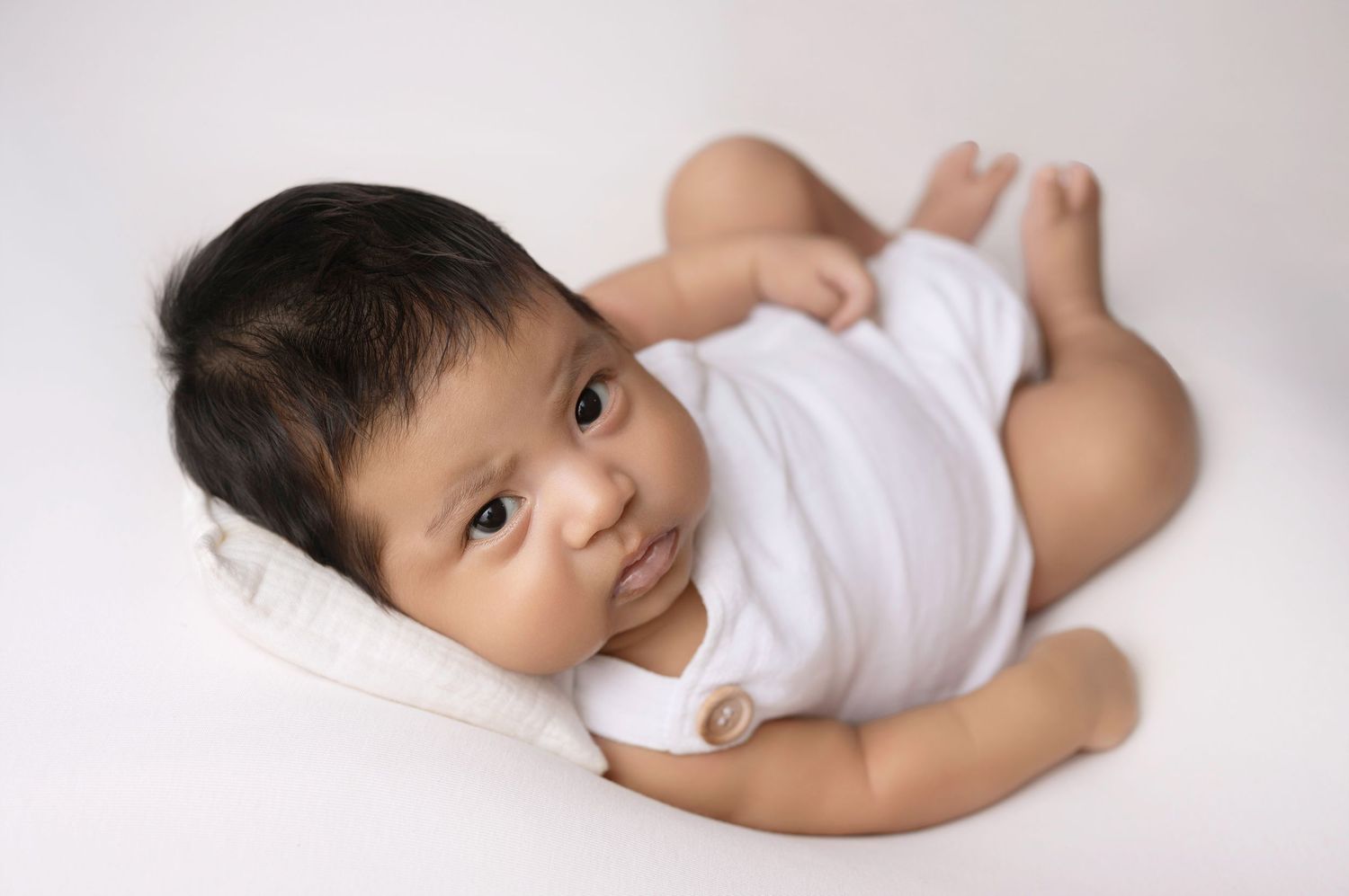 Adorable baby in white onesie lying on white background, looking up with dark hair and bright eyes.