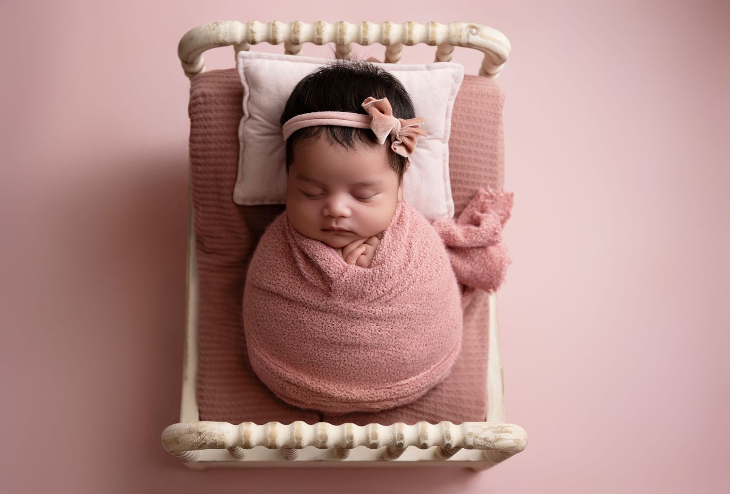 Newborn baby swaddled in pink wrap sleeping on miniature bed with bow headband and pink background.