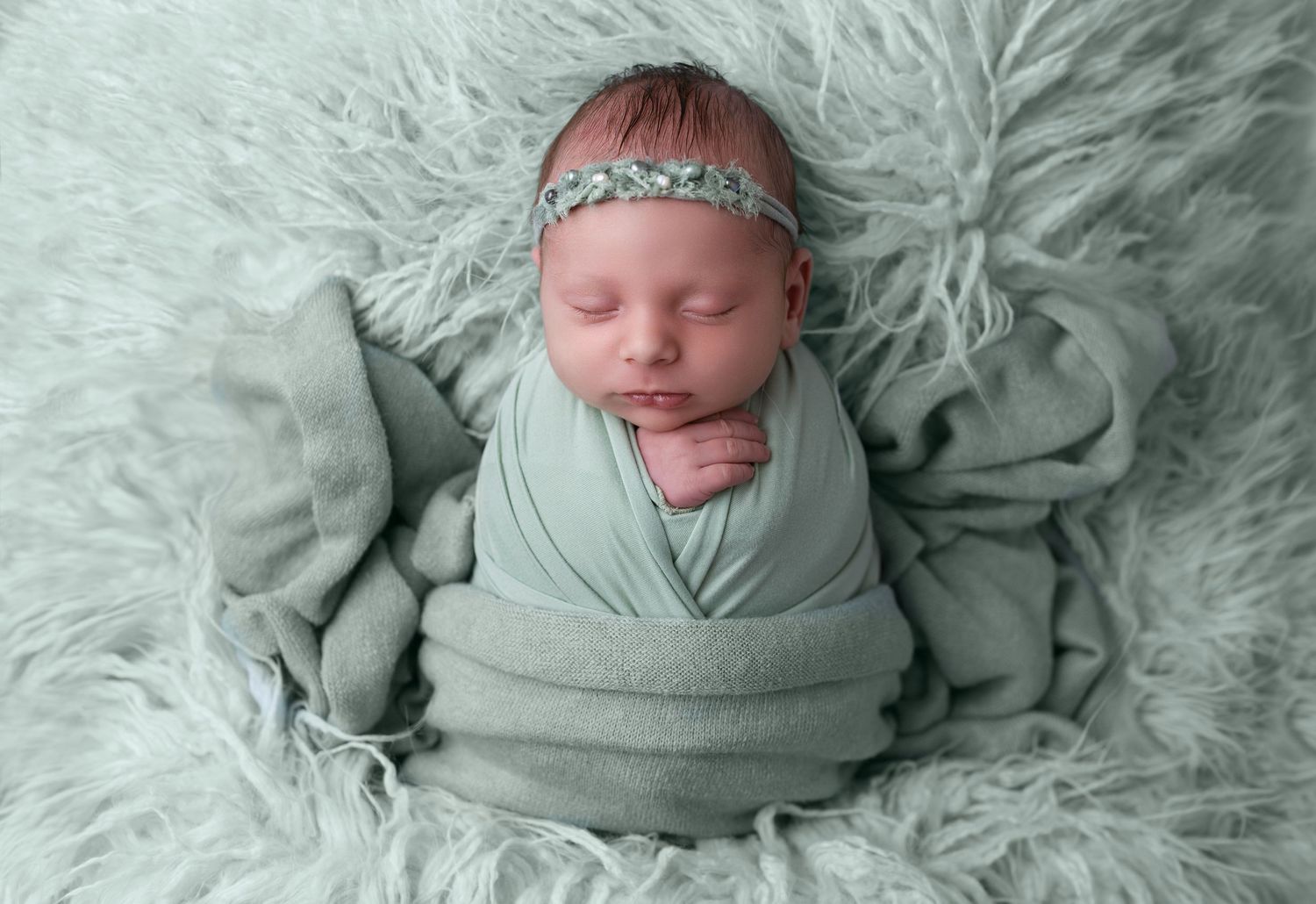 Sleeping newborn baby swaddled in sage green wrap on matching fluffy fur blanket with floral headband.