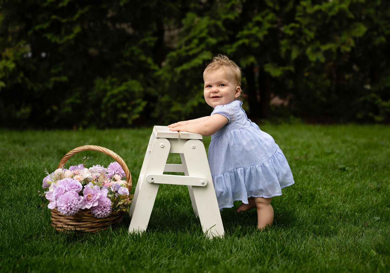 Toddler girl in blue dress standing by white step stool and flower basket on green grass outdoors.