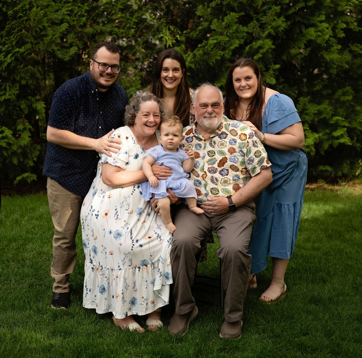 Multi-generational family portrait with six members posing outdoors on green grass, including a baby, smiling warmly.