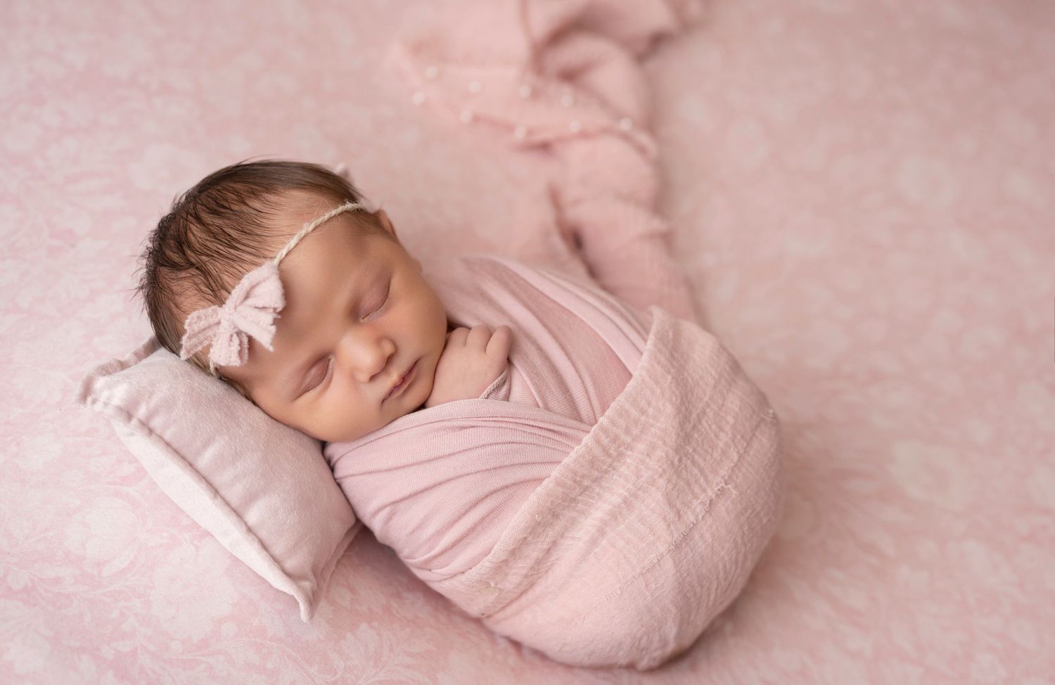 Newborn baby girl swaddled in pink wrap sleeping on pillow with bow headband on pink background.