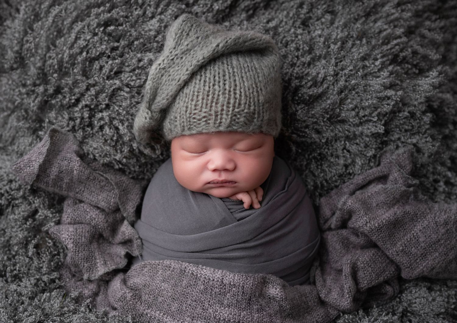 Newborn baby wrapped in gray swaddle wearing knit slouchy hat sleeping on gray fur blanket.