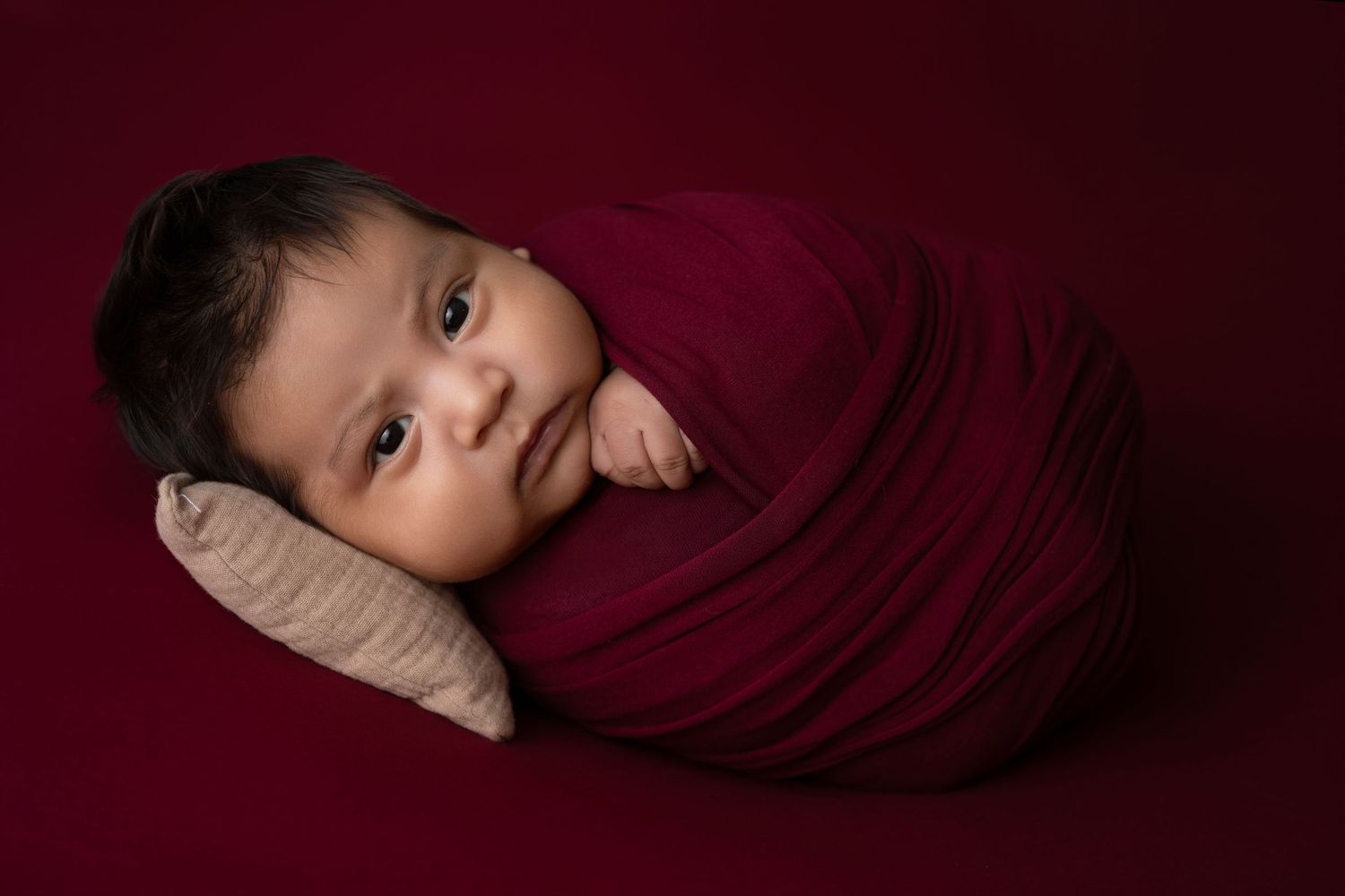 Newborn baby swaddled in a deep red wrap, resting on a pillow against a burgundy background.