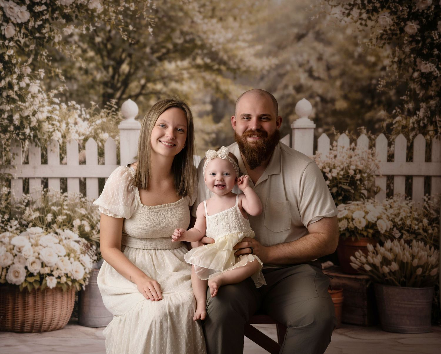 Family portrait of smiling parents with baby girl in yellow dress, posed before white picket fence and floral backdrop.