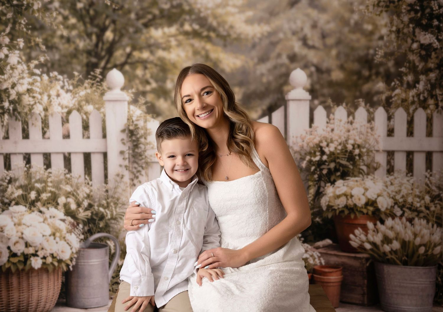 A woman in a white dress poses with a young boy in a white shirt against a floral garden backdrop.