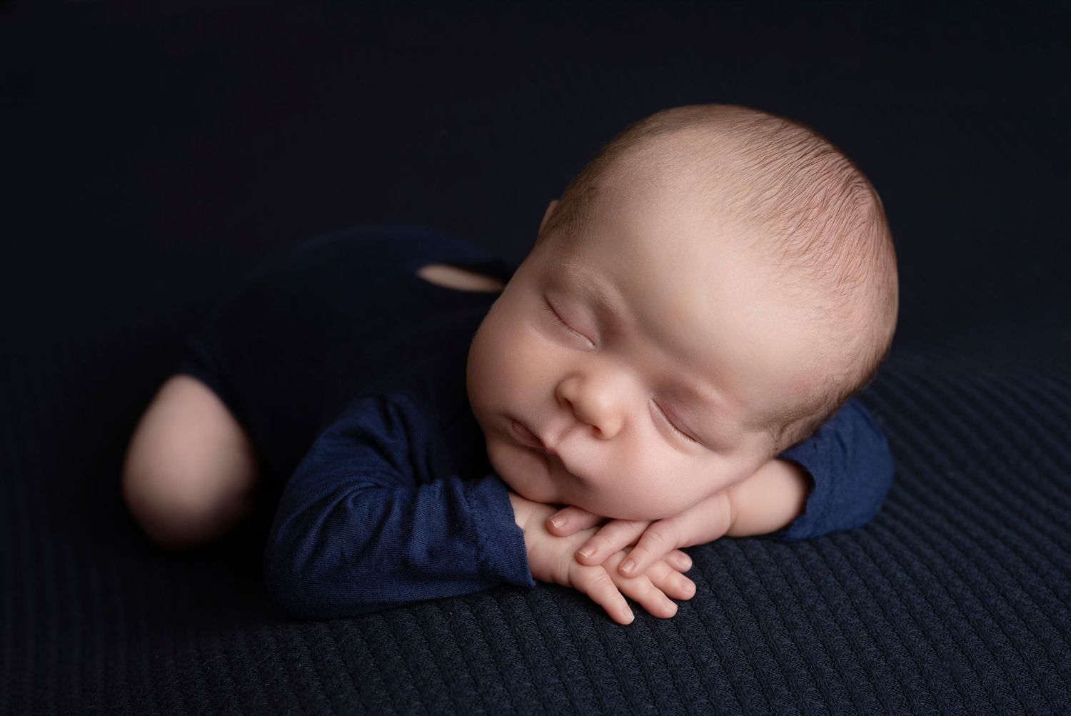 Peaceful newborn baby sleeping in froggy pose wearing navy blue outfit on dark textured blanket.