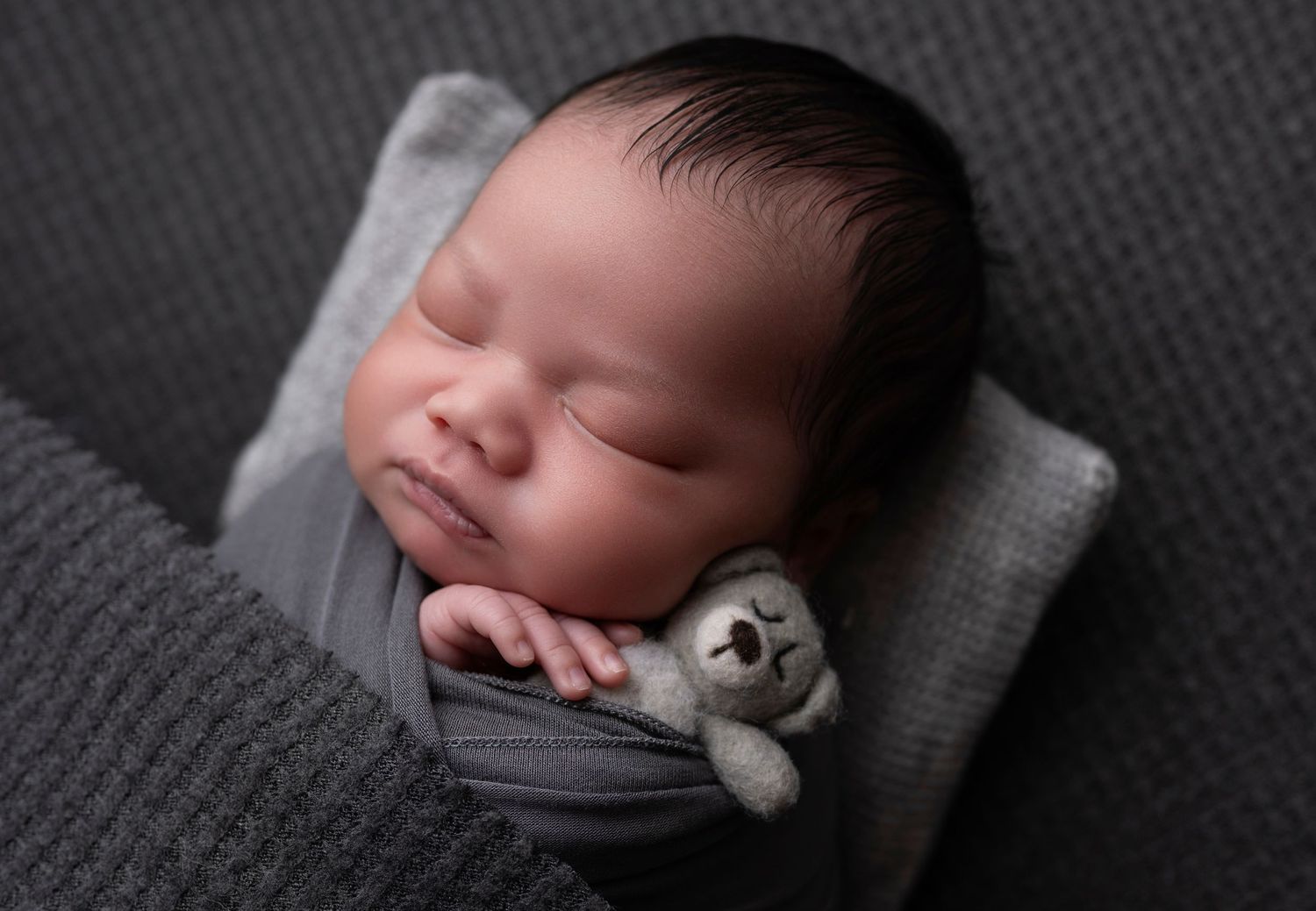 Sleeping newborn baby in gray wrap cuddling small teddy bear on gray textured pillow.