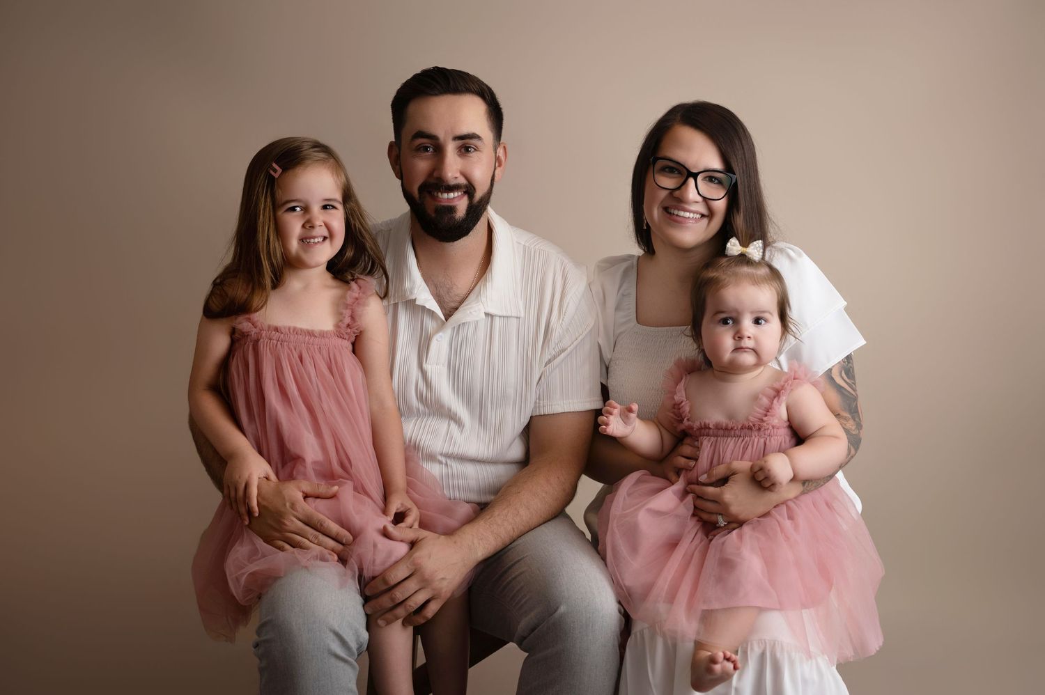 Family portrait of parents with two young daughters in matching pink dresses against a neutral beige background.