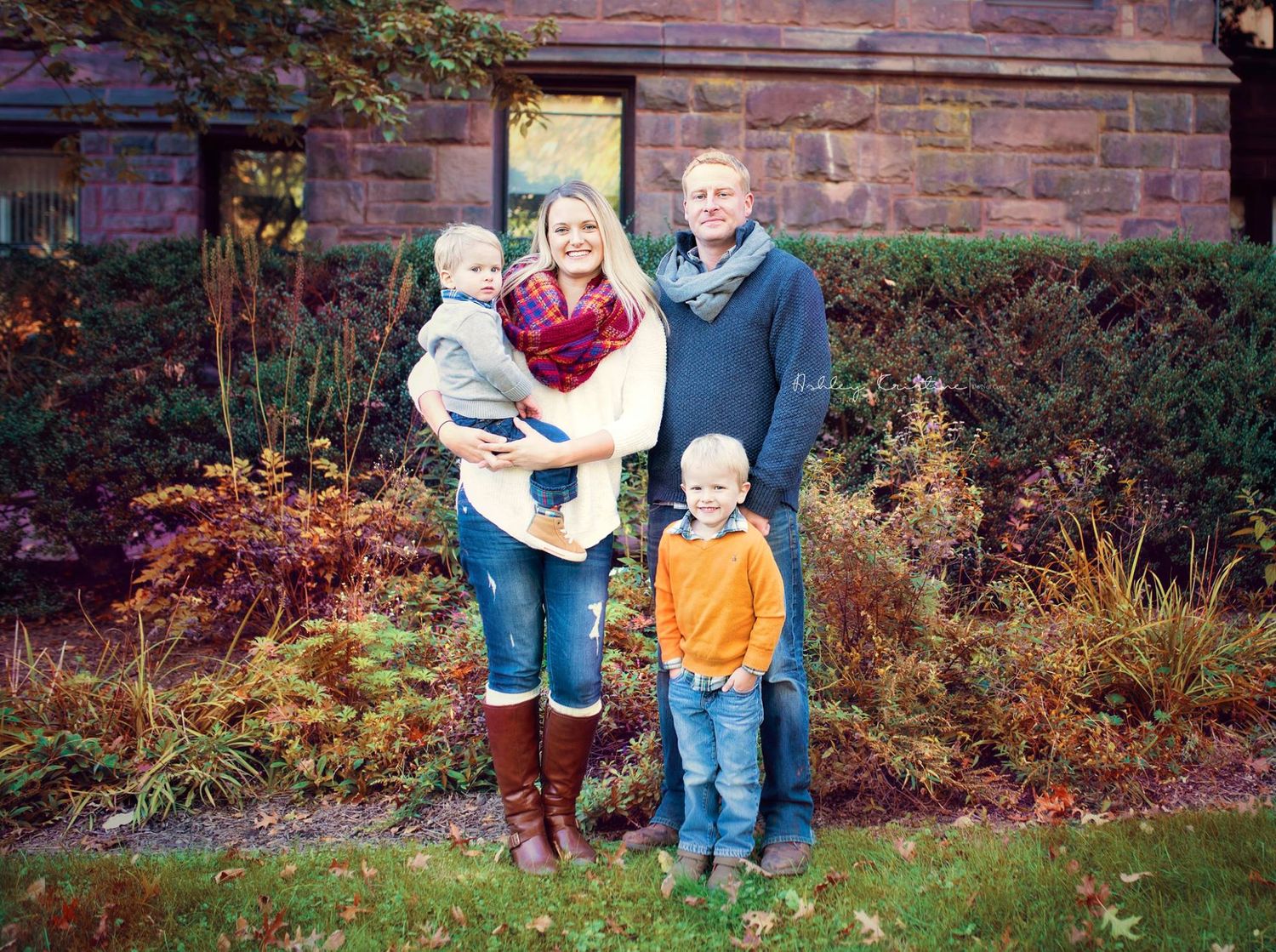 Family of four posing outdoors in front of stone building with fall foliage.