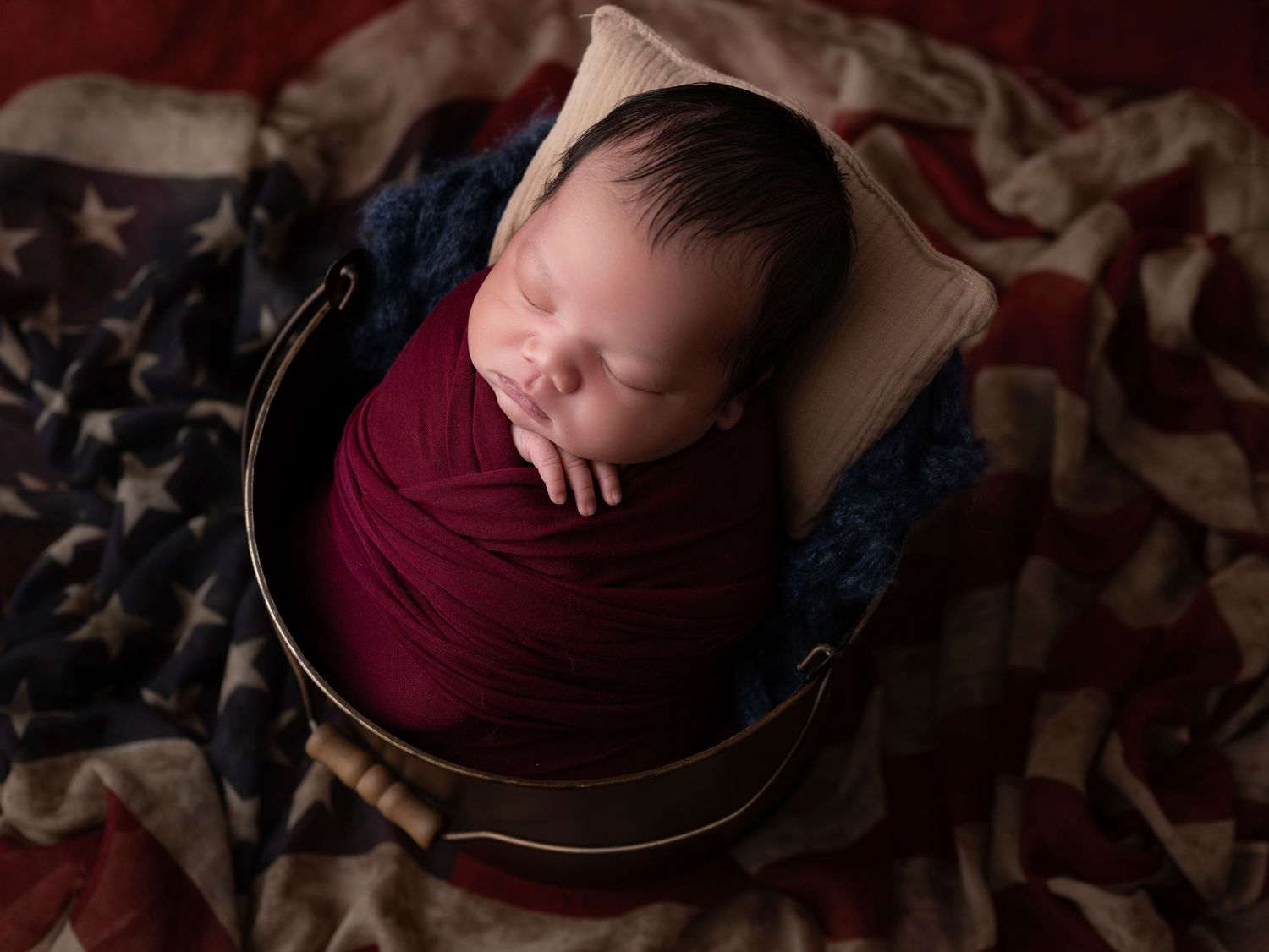 Newborn baby wrapped in burgundy swaddle sleeping in bucket on American flag patriotic backdrop.