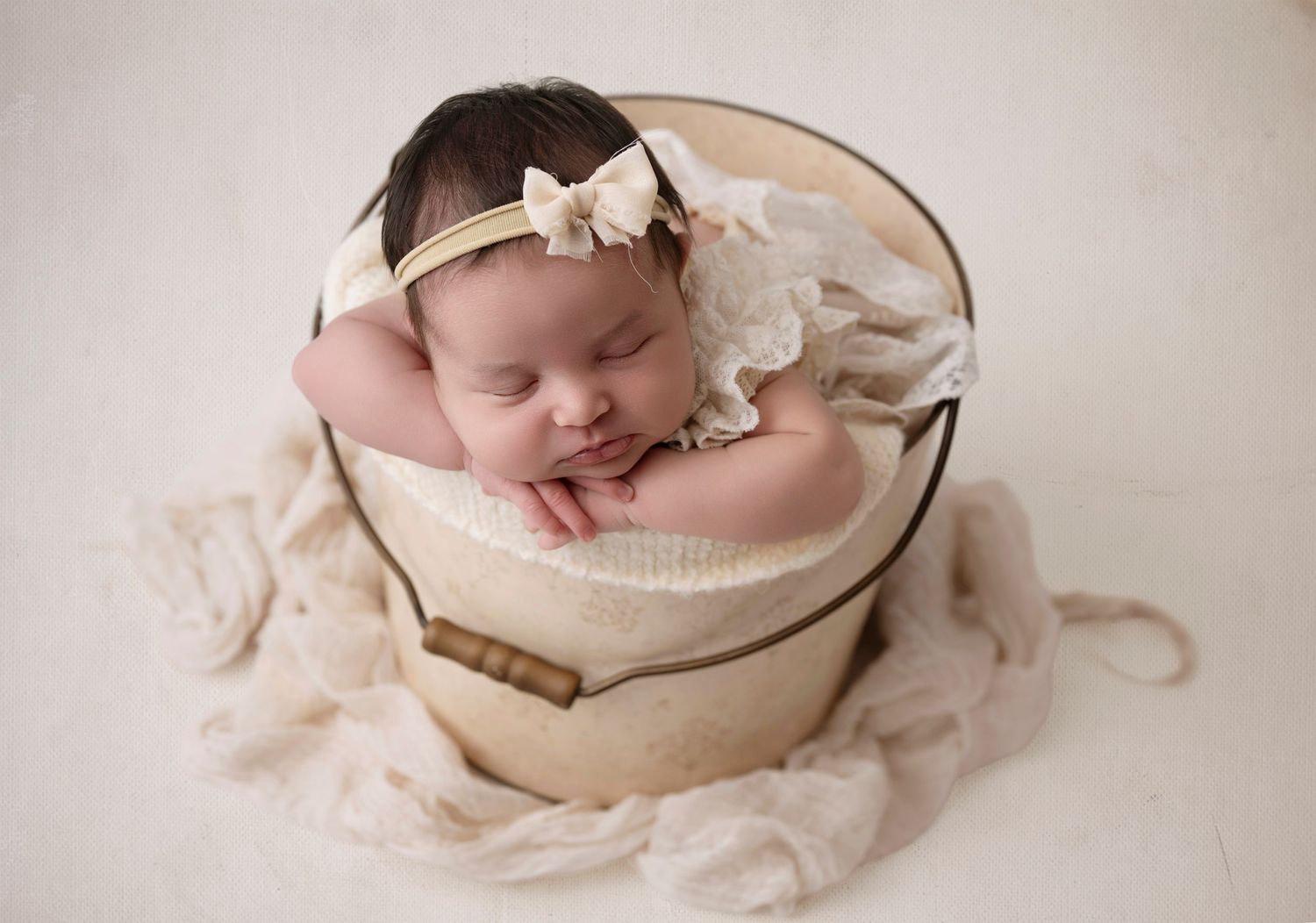 Peaceful newborn baby sleeping in a wooden bucket with cream lace wrap and bow headband.