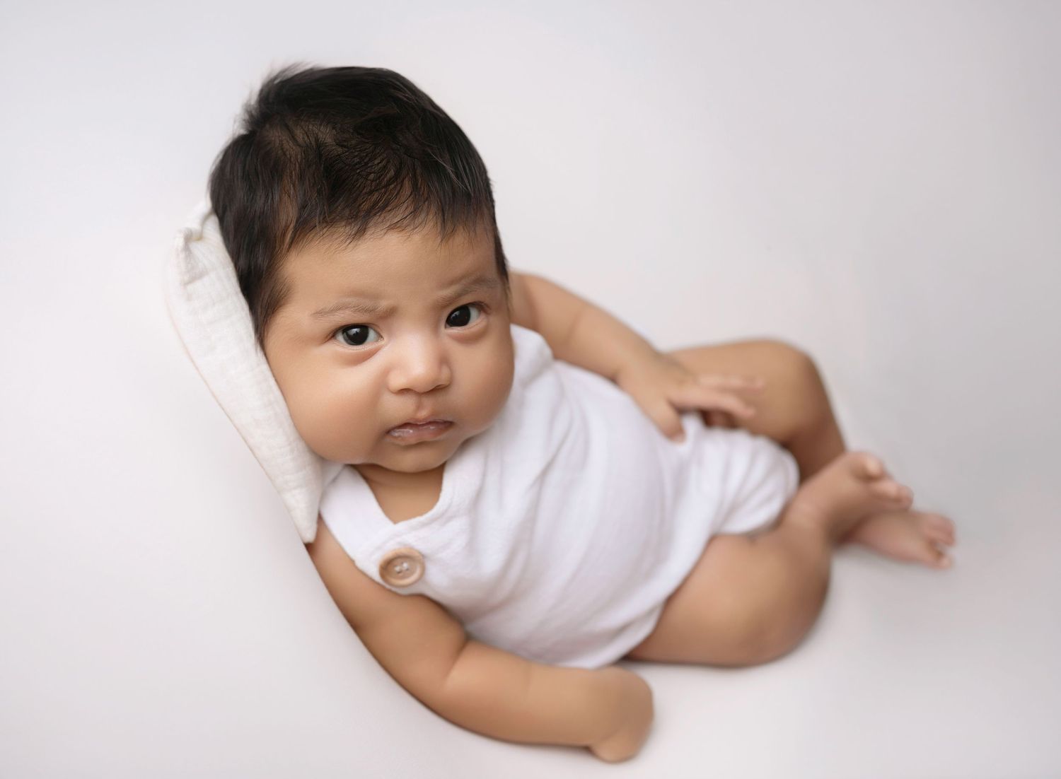 Alert dark-haired baby in white onesie resting on pillow against clean white background. Newborn photoshoot