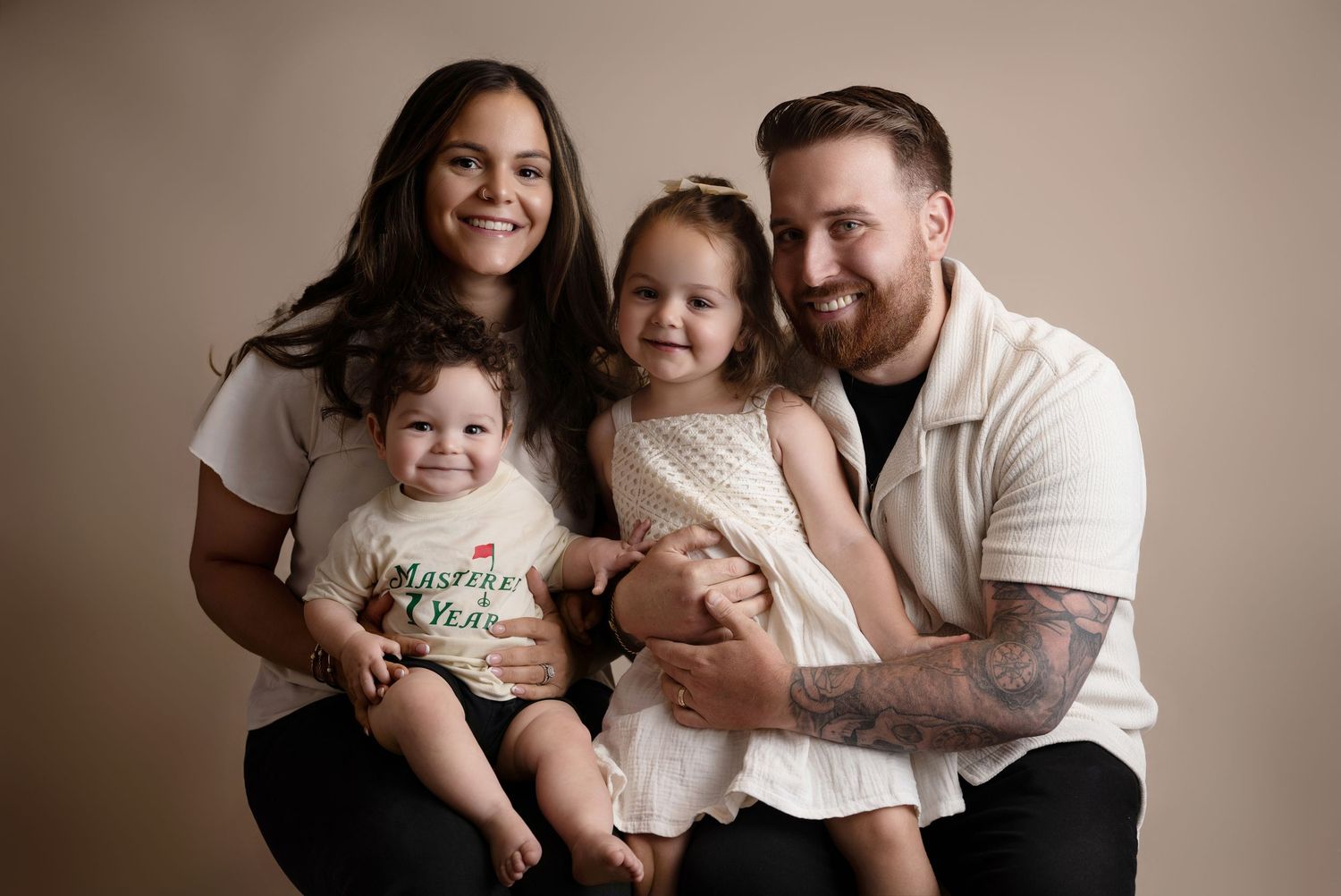 Happy family of four posing together, parents holding two young children, smiling against a neutral beige background.