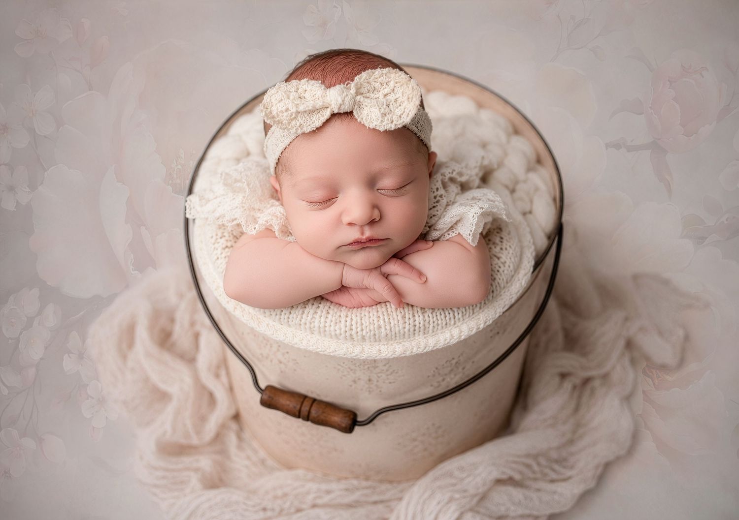 Peaceful newborn baby posed in cream bucket with floral headband on soft textured backdrop.
