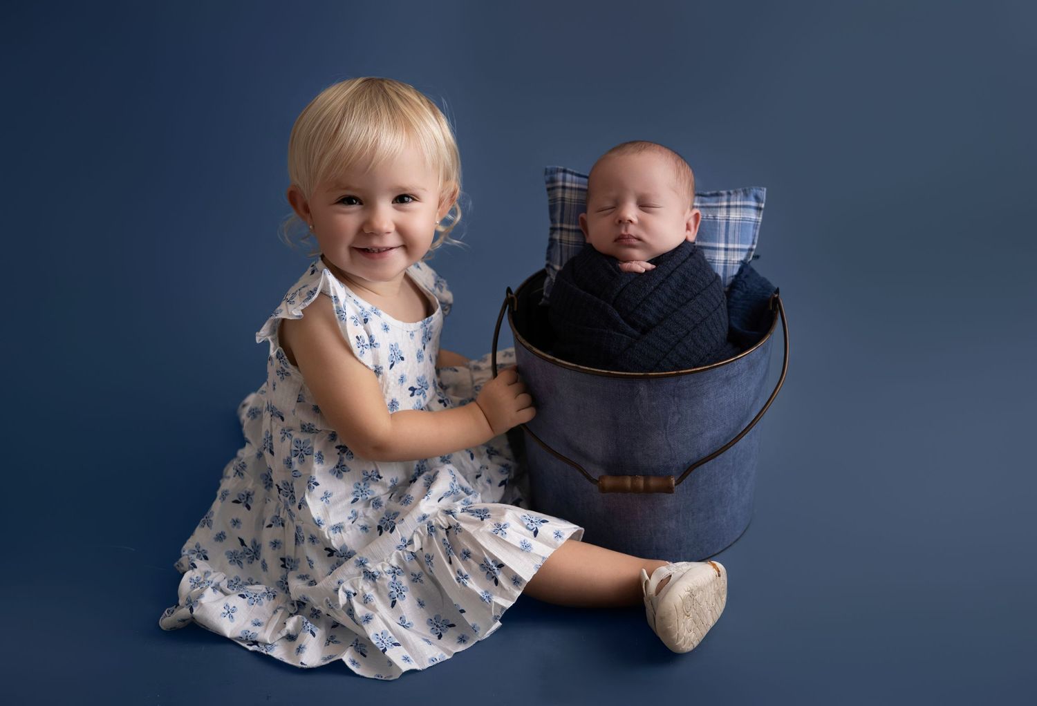 Toddler girl in floral dress smiles beside newborn baby sleeping in decorative blue bucket on blue background.