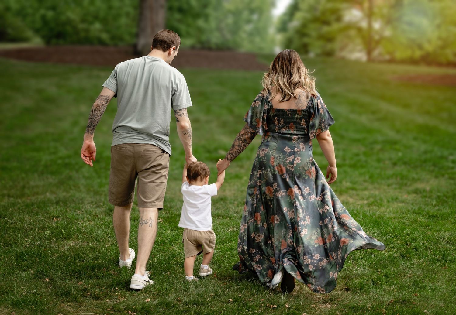 Young family walking through green park holding hands with toddler between parents from behind.