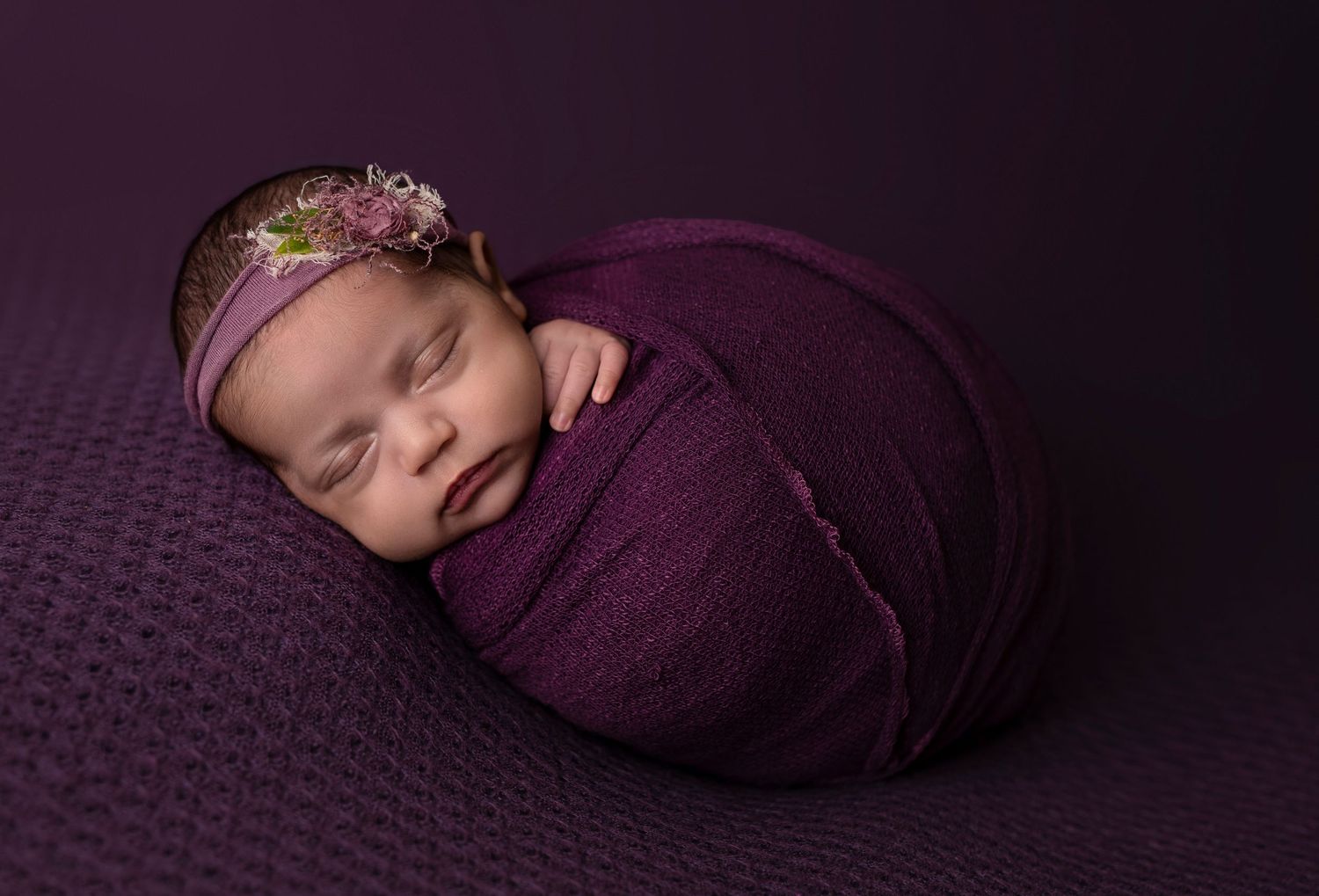 Sleeping newborn wrapped in purple swaddle with floral headband on purple backdrop.