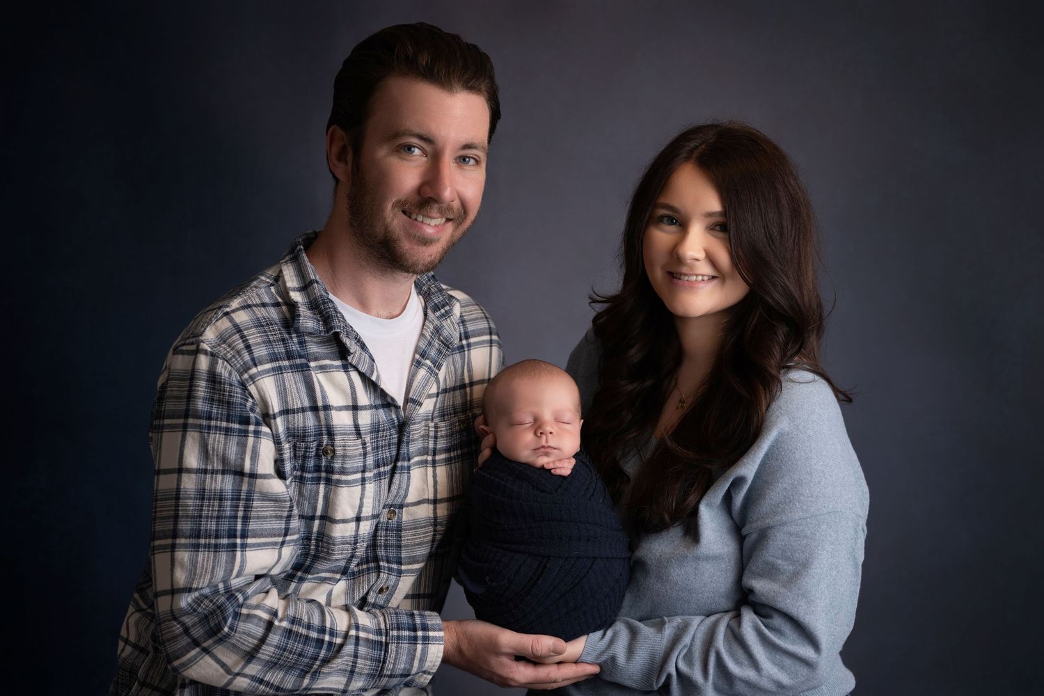 A couple smiles holding their swaddled newborn baby in a professional studio portrait against a dark background.