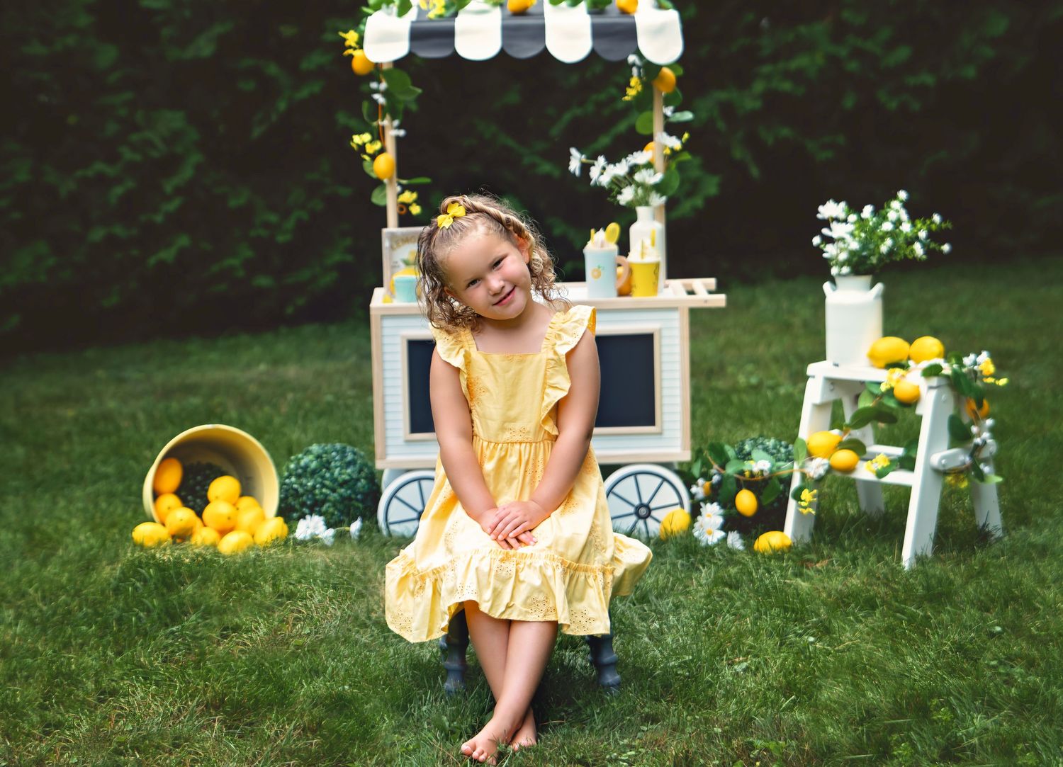 Smiling young girl in yellow dress sits beside a lemonade stand cart surrounded by lemons and flowers.