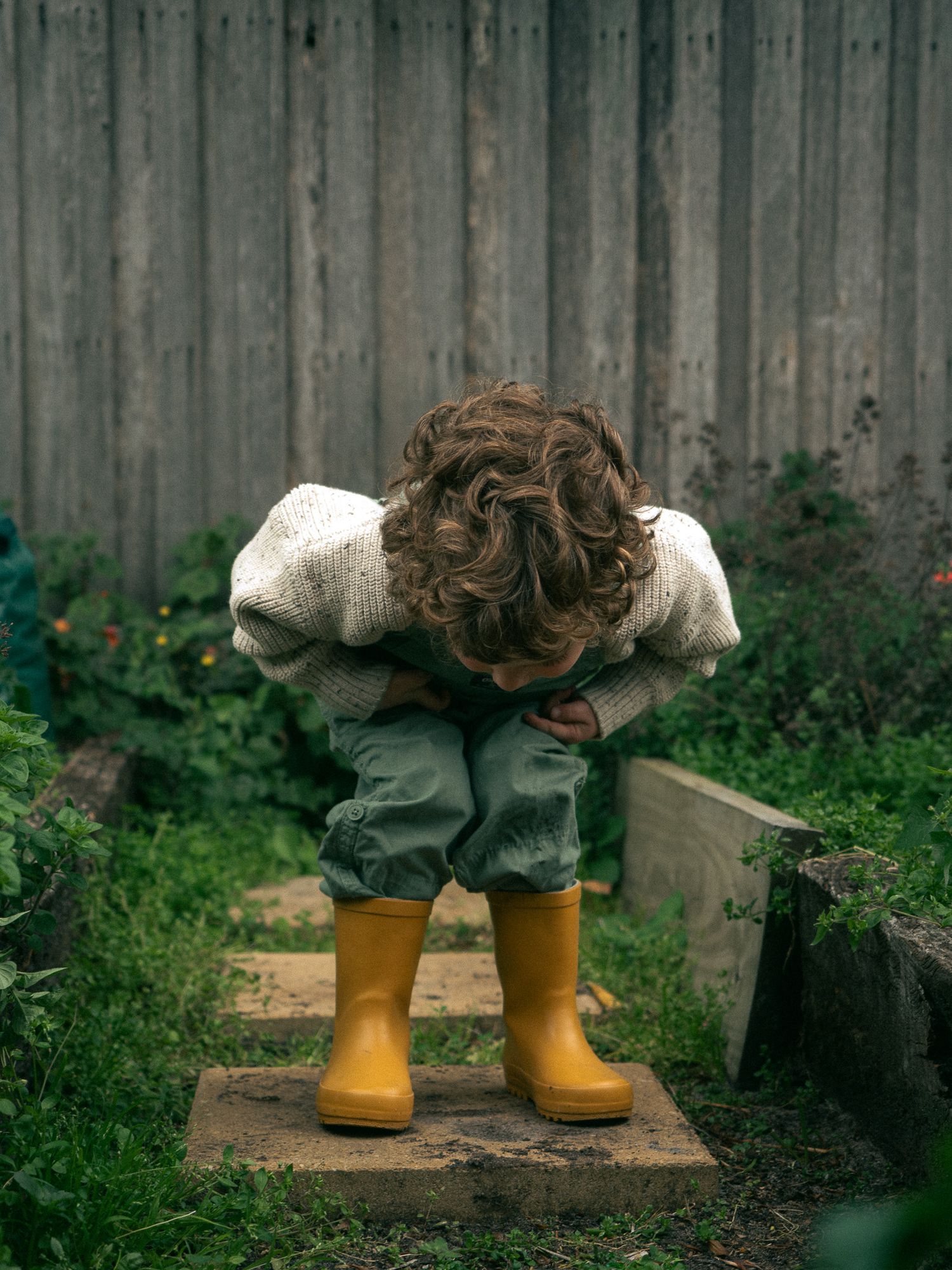 A boy in yellow rain boots standing in a garden against a wooden fence with greenery in the background.