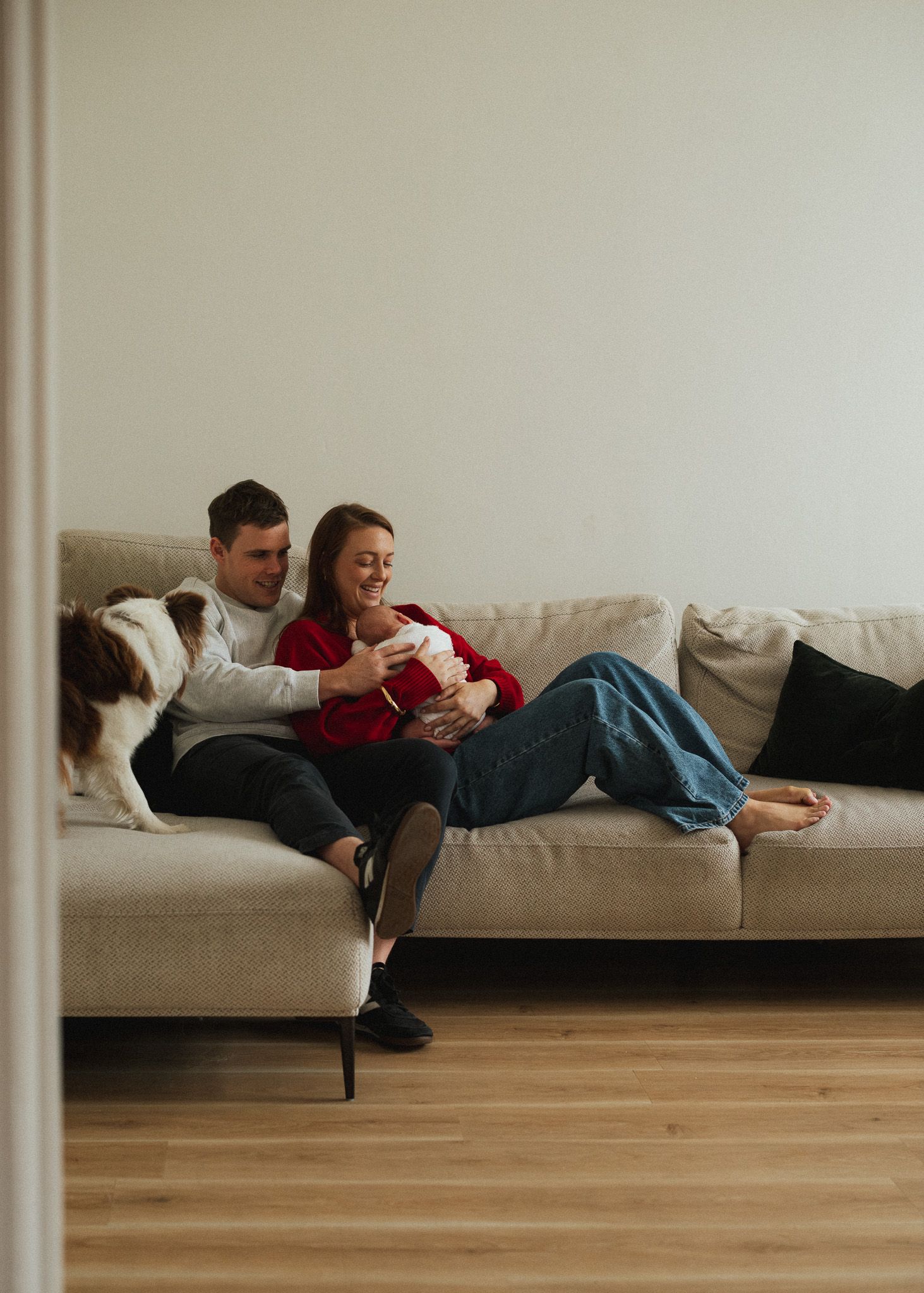 Happy couple relaxing on a beige sofa with their dog, holding a newborn baby in a cozy modern living room.