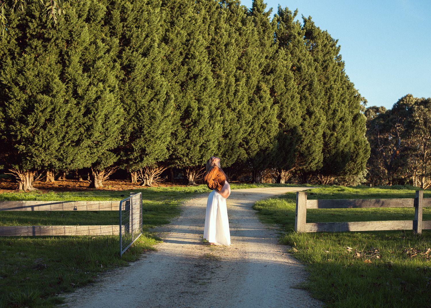 Woman in white dress standing on a rural gravel path flanked by wooden fences and tall green trees.