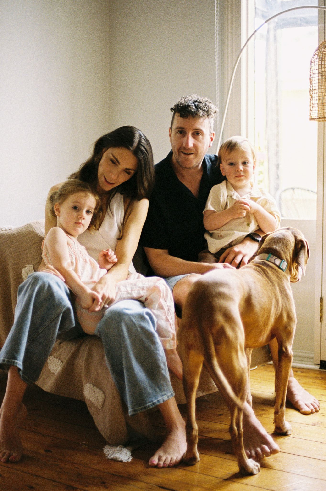 A family relaxes together on a couch in a sunlit room with their brown dog standing beside them.