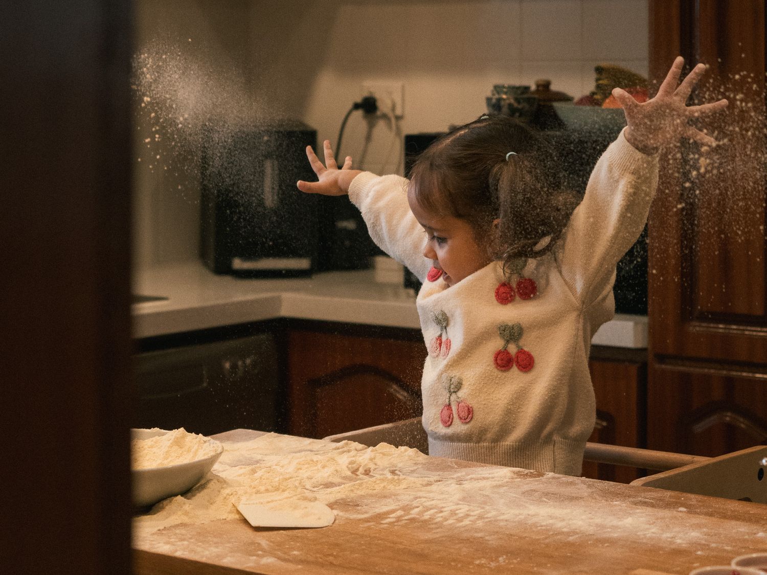 Young child joyfully tossing flour in a kitchen while baking, wearing a cherry-print sweater.
