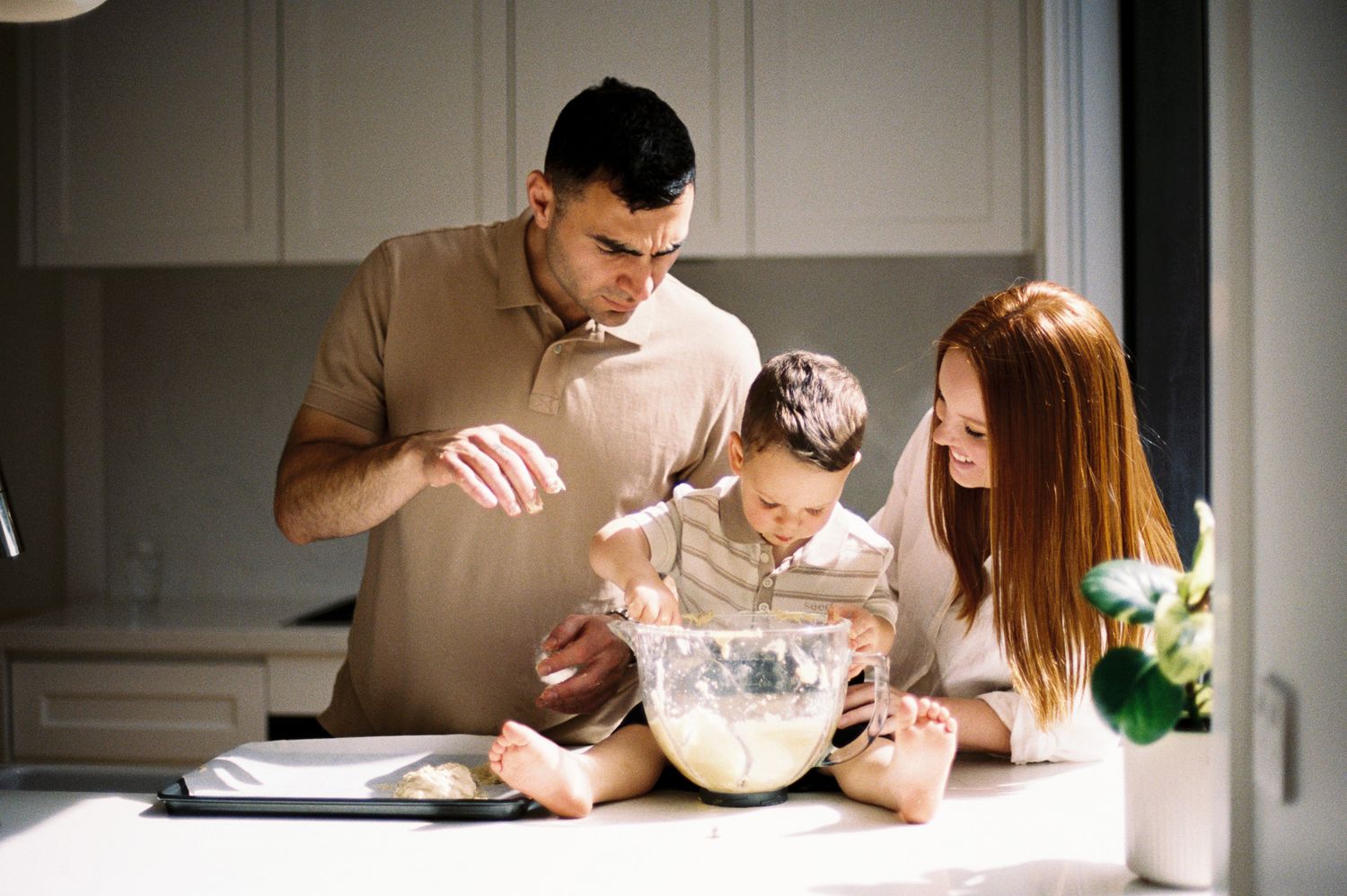 A family bakes together in their kitchen, mixing ingredients in a large white bowl on the counter.