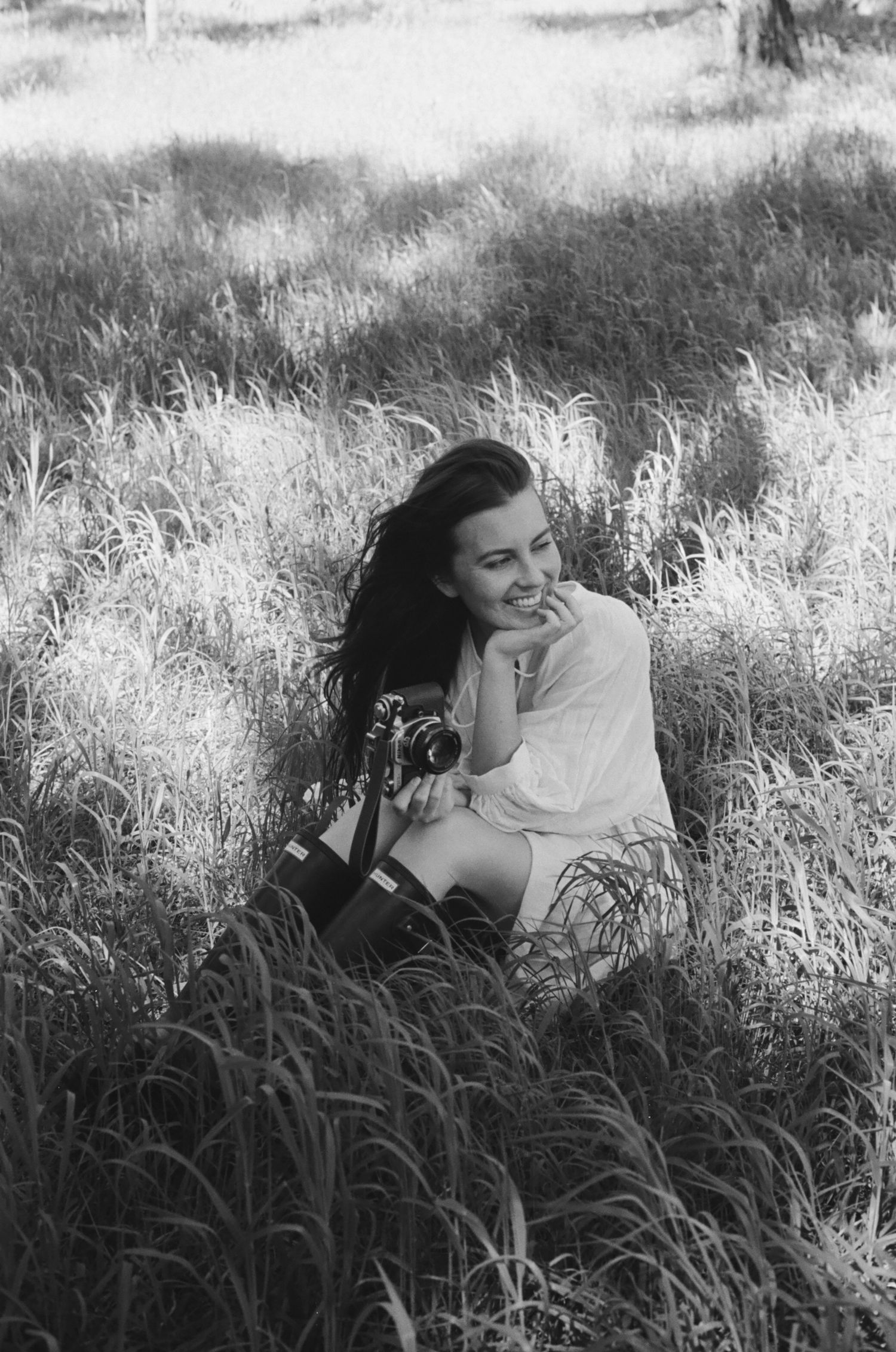 Black and white photo of a smiling woman sitting in tall grass holding a vintage camera outdoors.