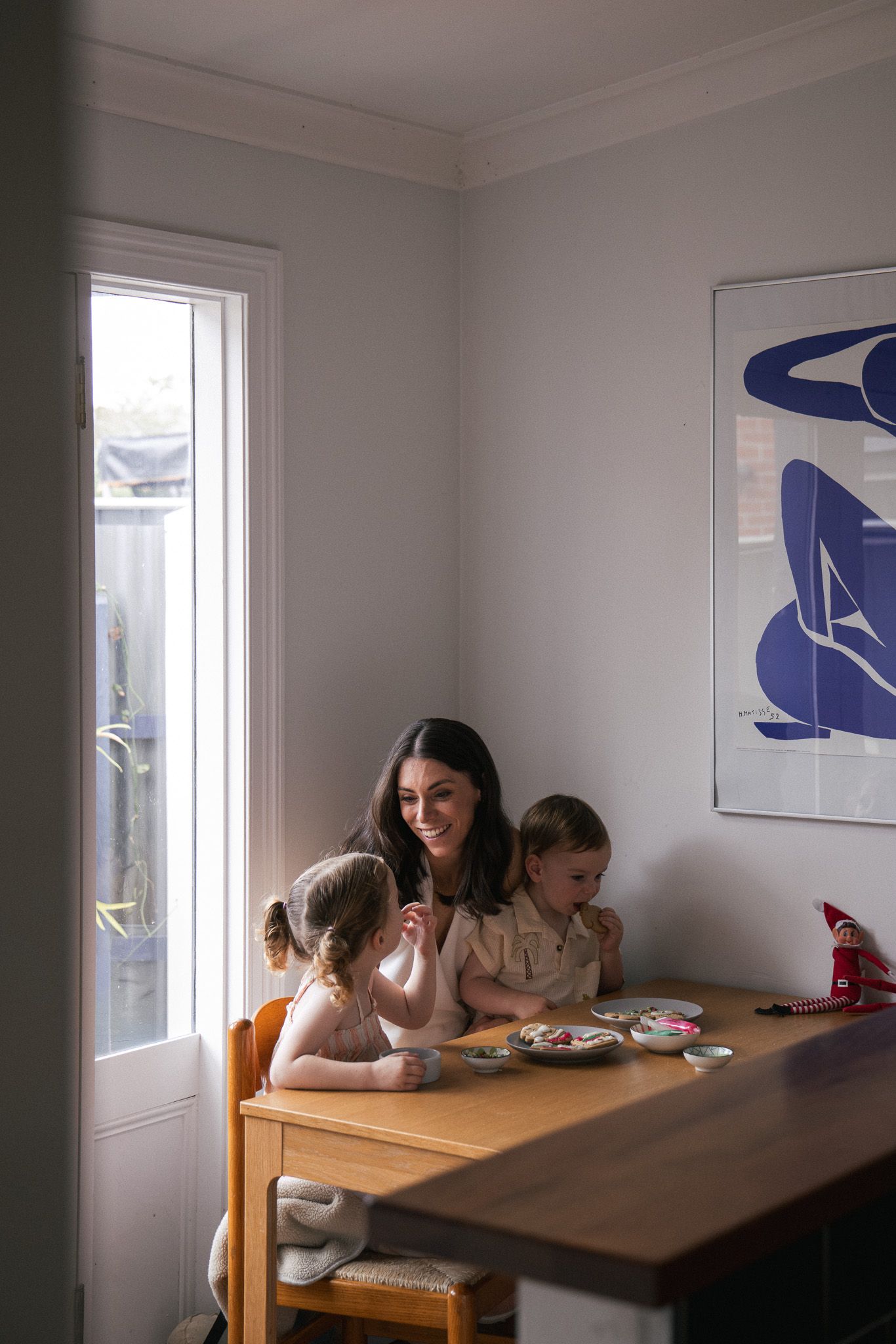 A smiling mother sits with two young children at a wooden dining table near a bright window in a modern home.