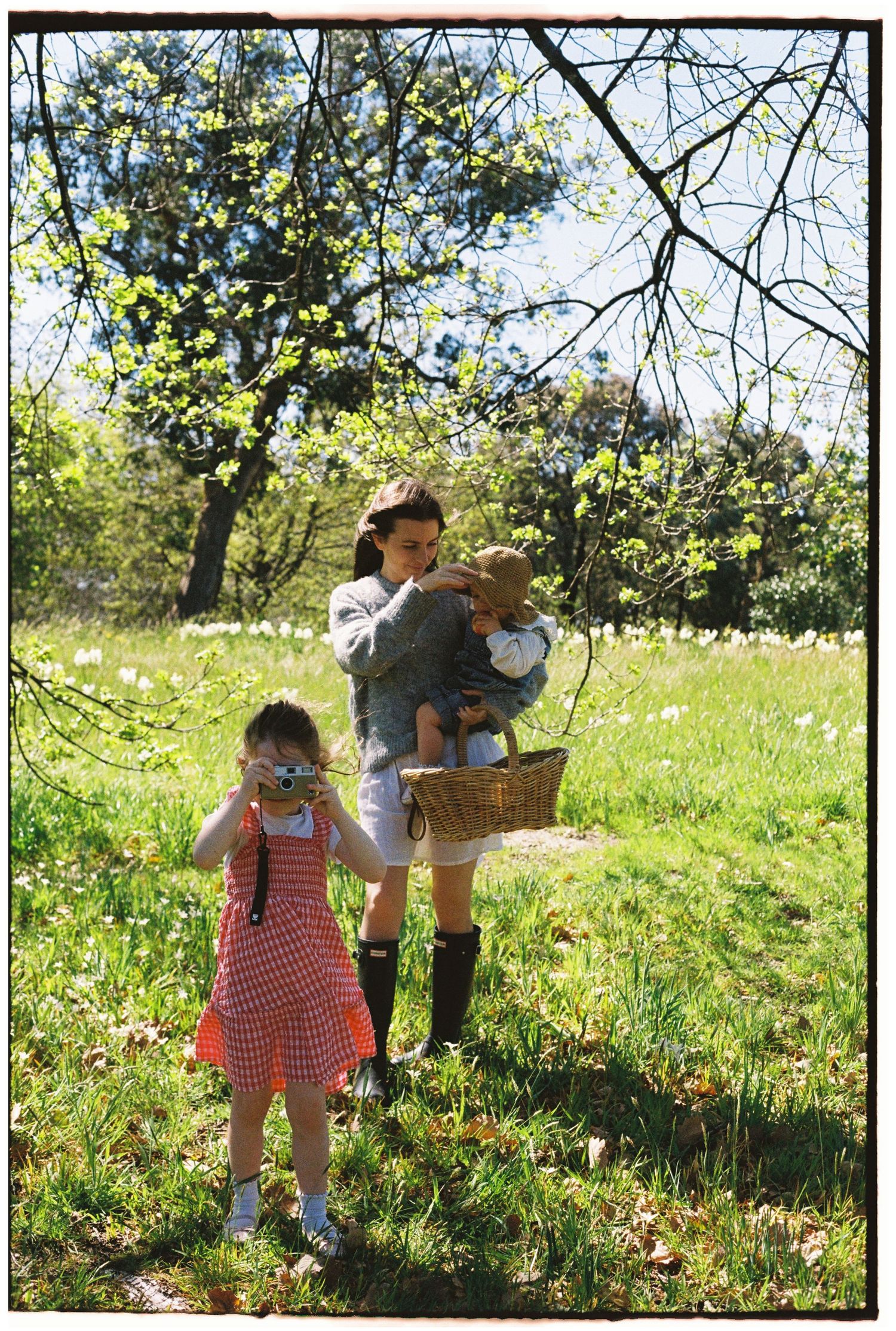 A woman holding a baby and basket stands in a sunny meadow while a young girl in a red dress photographs with a camera.