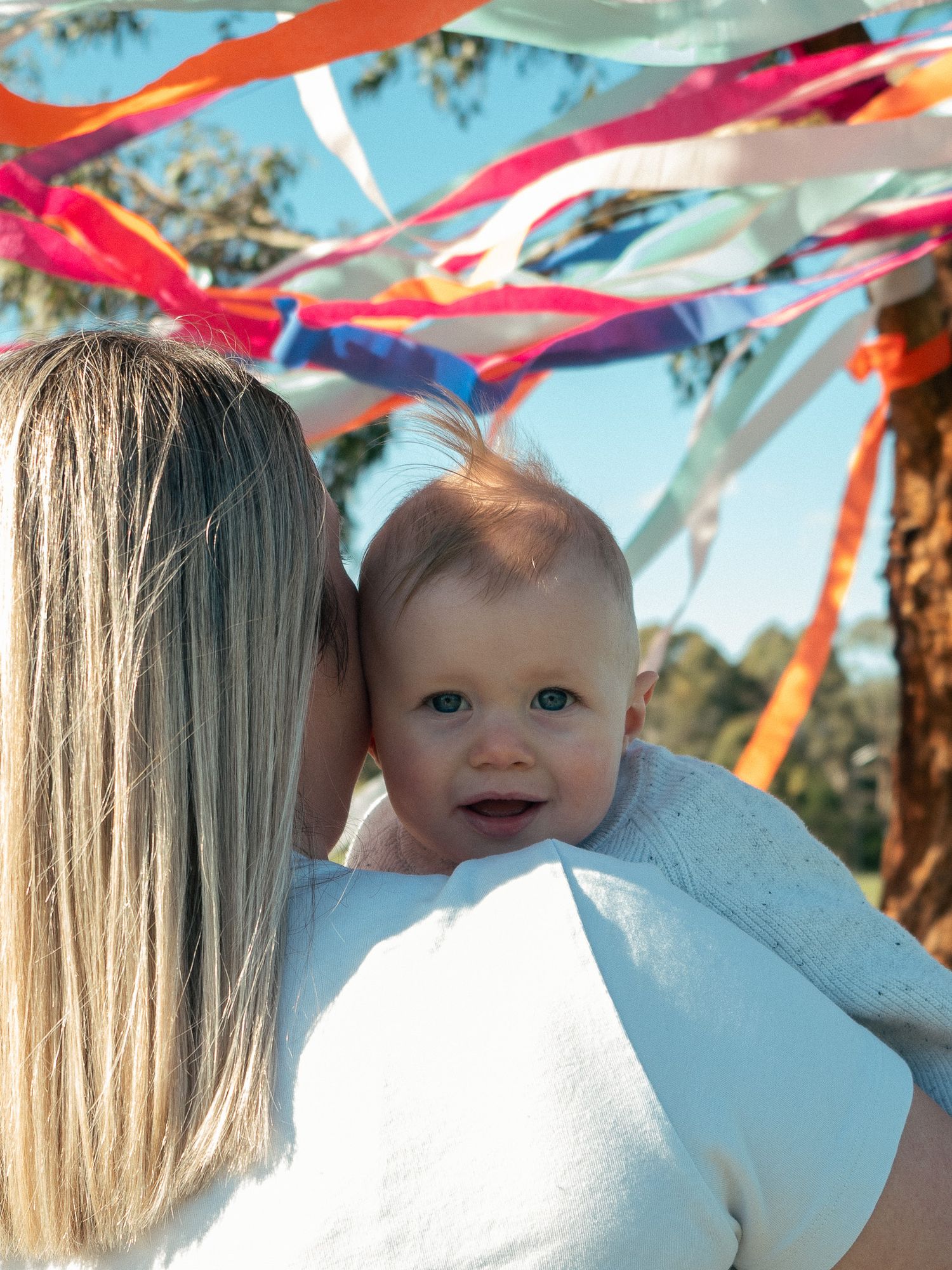 Colorful streamers hanging from tree branches for a motherhood session with the infant looking over the mum's shoulder looking at the camera