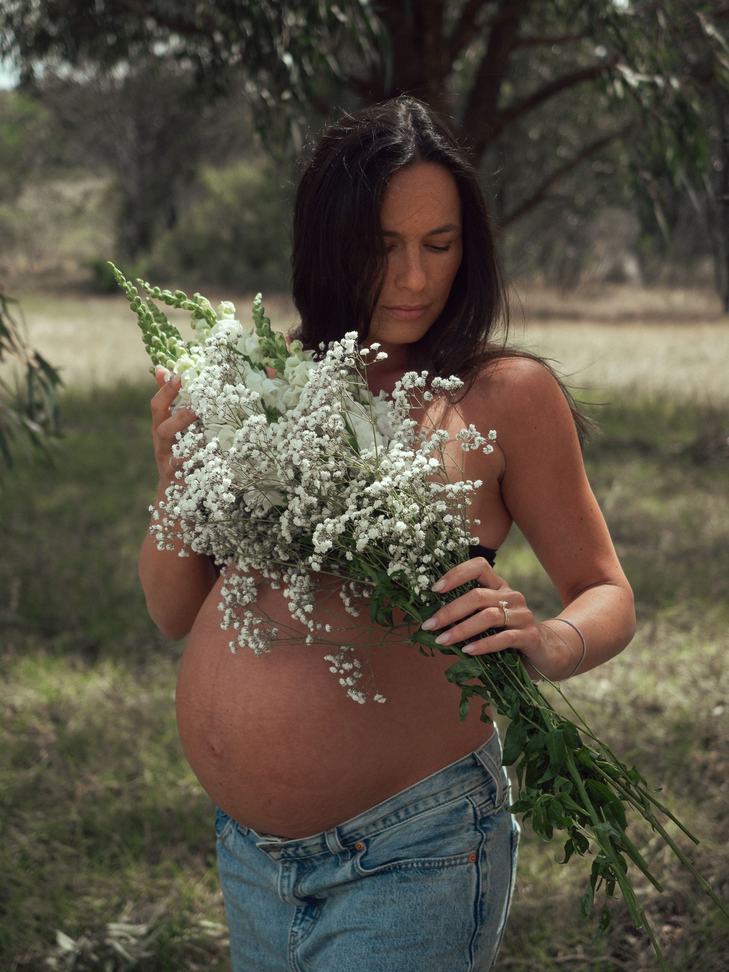 A pregnant person in jeans holds a delicate bouquet of white baby's breath flowers against their bare belly in nature.