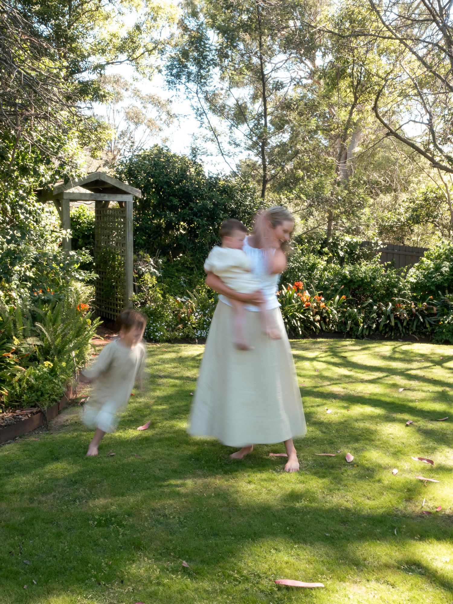 Children in white dresses spin and dance playfully together on a sunny lawn surrounded by trees and greenery.