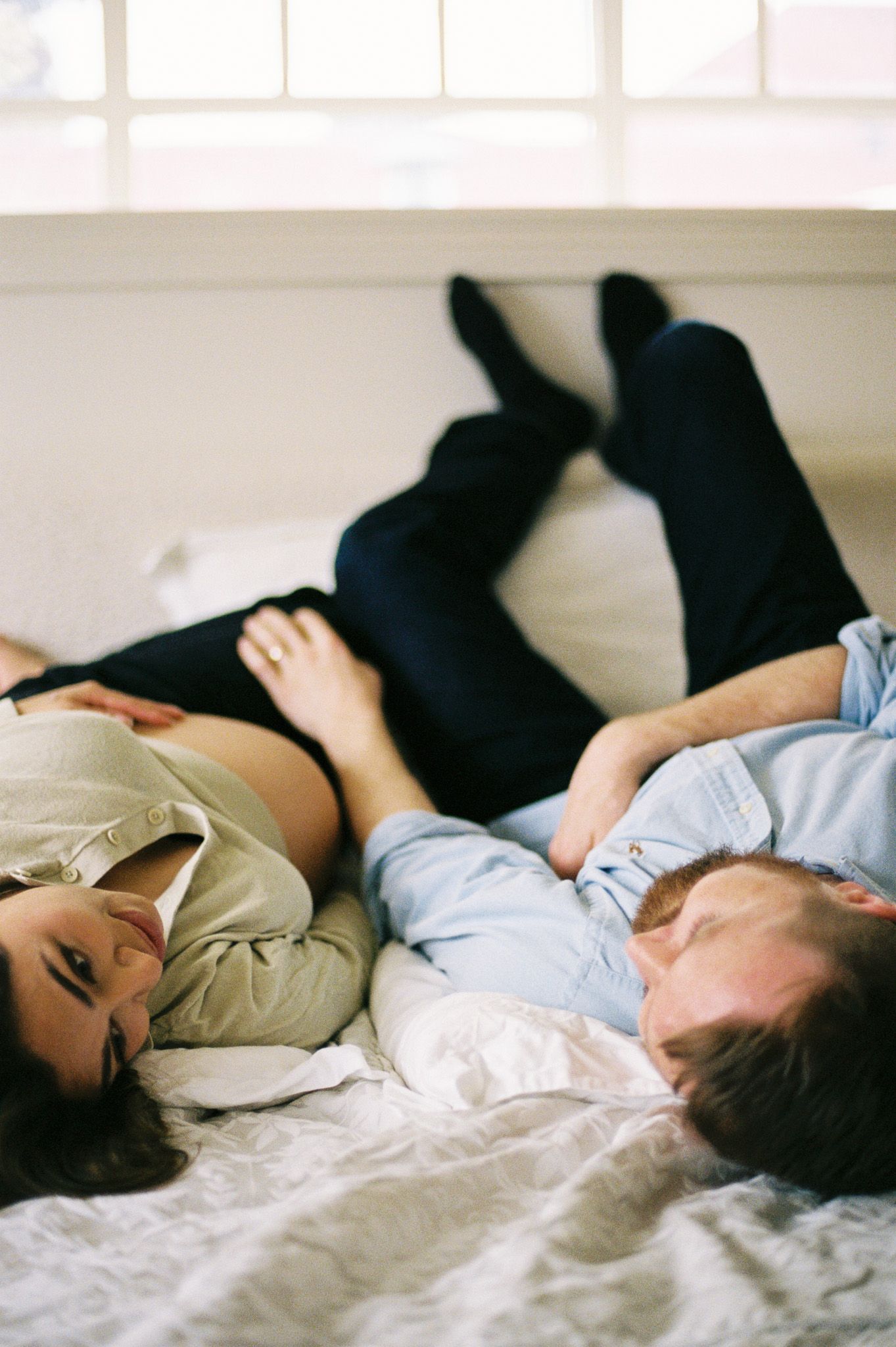 A sequence of a couple during their in-home maternity session peaceful resting on white bedding in natural light.