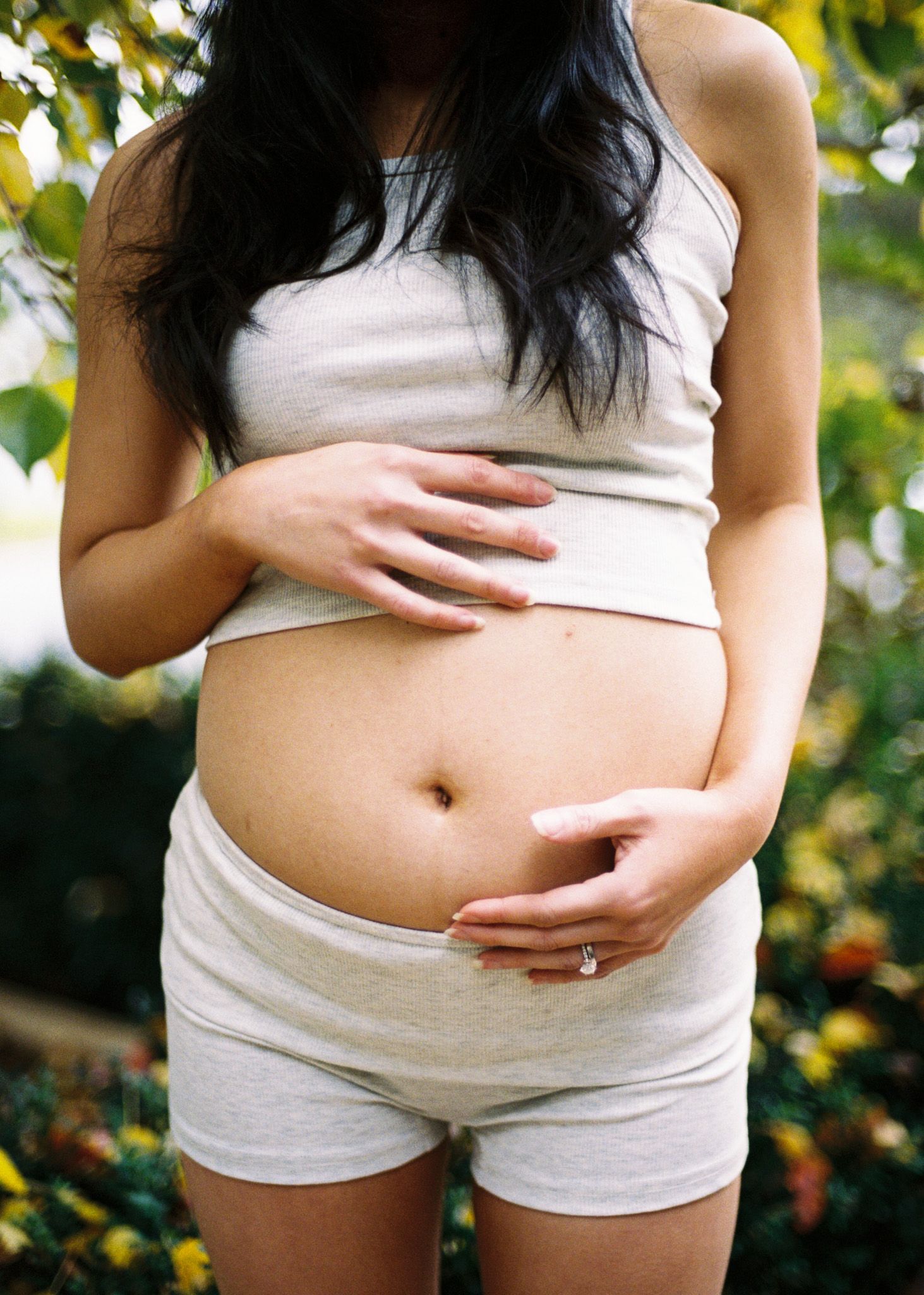 Pregnant woman in white crop top and shorts cradling baby bump outdoors among lush green foliage.