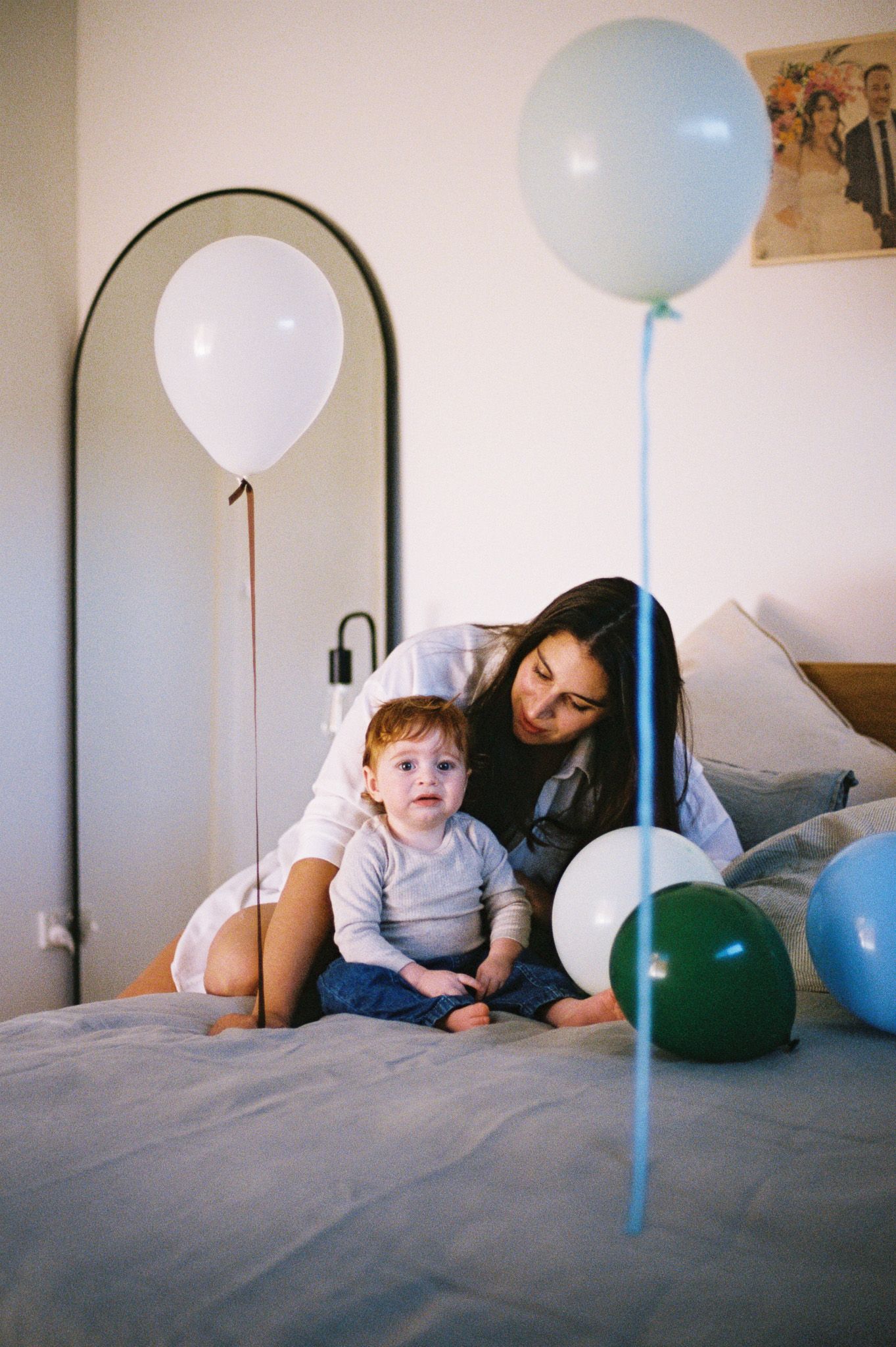 A mother and toddler sit together on a bed surrounded by colorful balloons during a birthday celebration.