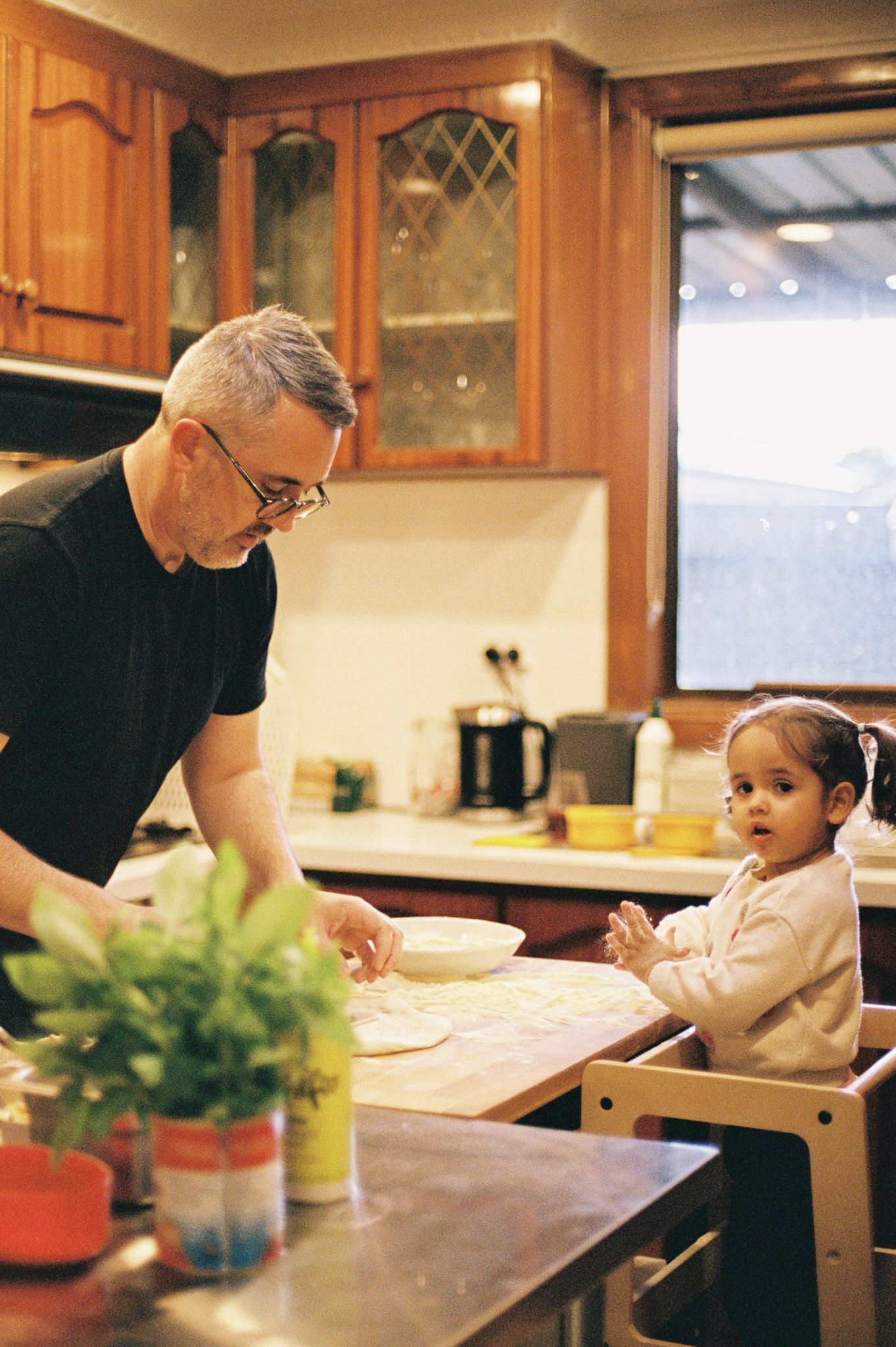 Man in black shirt rolling dough at kitchen counter while toddler watches from high chair in warm wooden kitchen.
