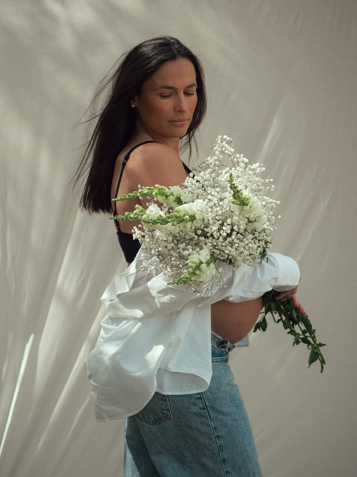 A woman in a white blouse and jeans holds a delicate bouquet of white baby's breath flowers against a light backdrop.
