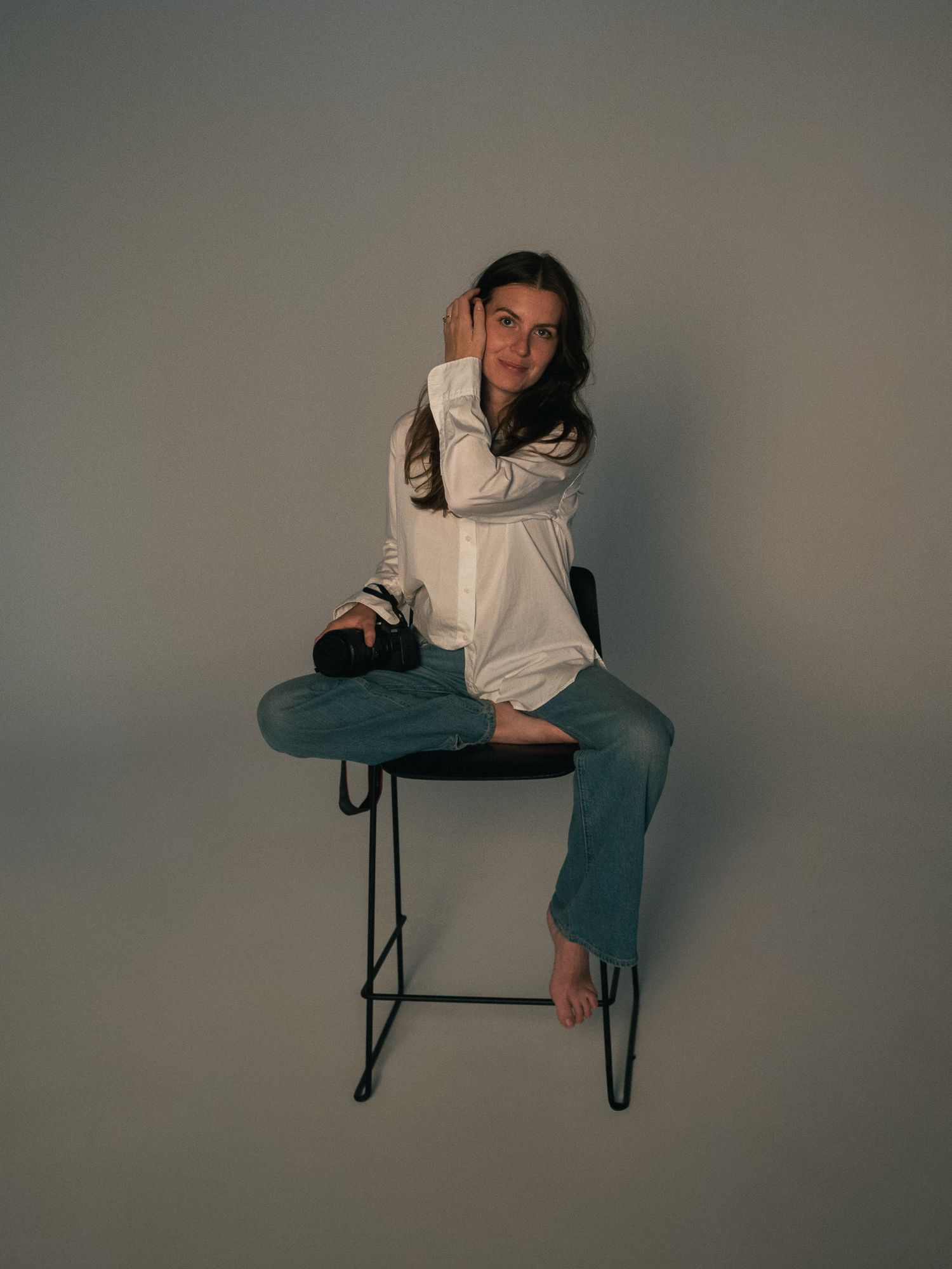 A person in a beige sweater and blue jeans sits casually on a tall stool against a gray studio backdrop.